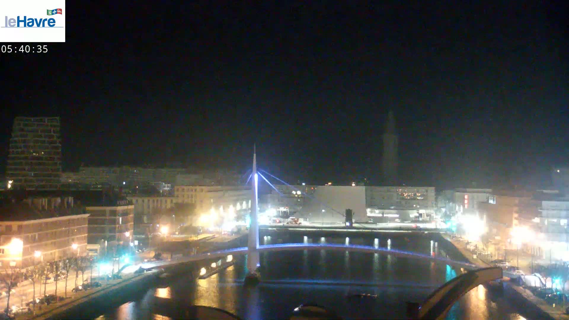 A nighttime view of a city waterfront, showing a lit bridge spanning a canal, with buildings and streetlights illuminated along the shore under a clear, dark sky.