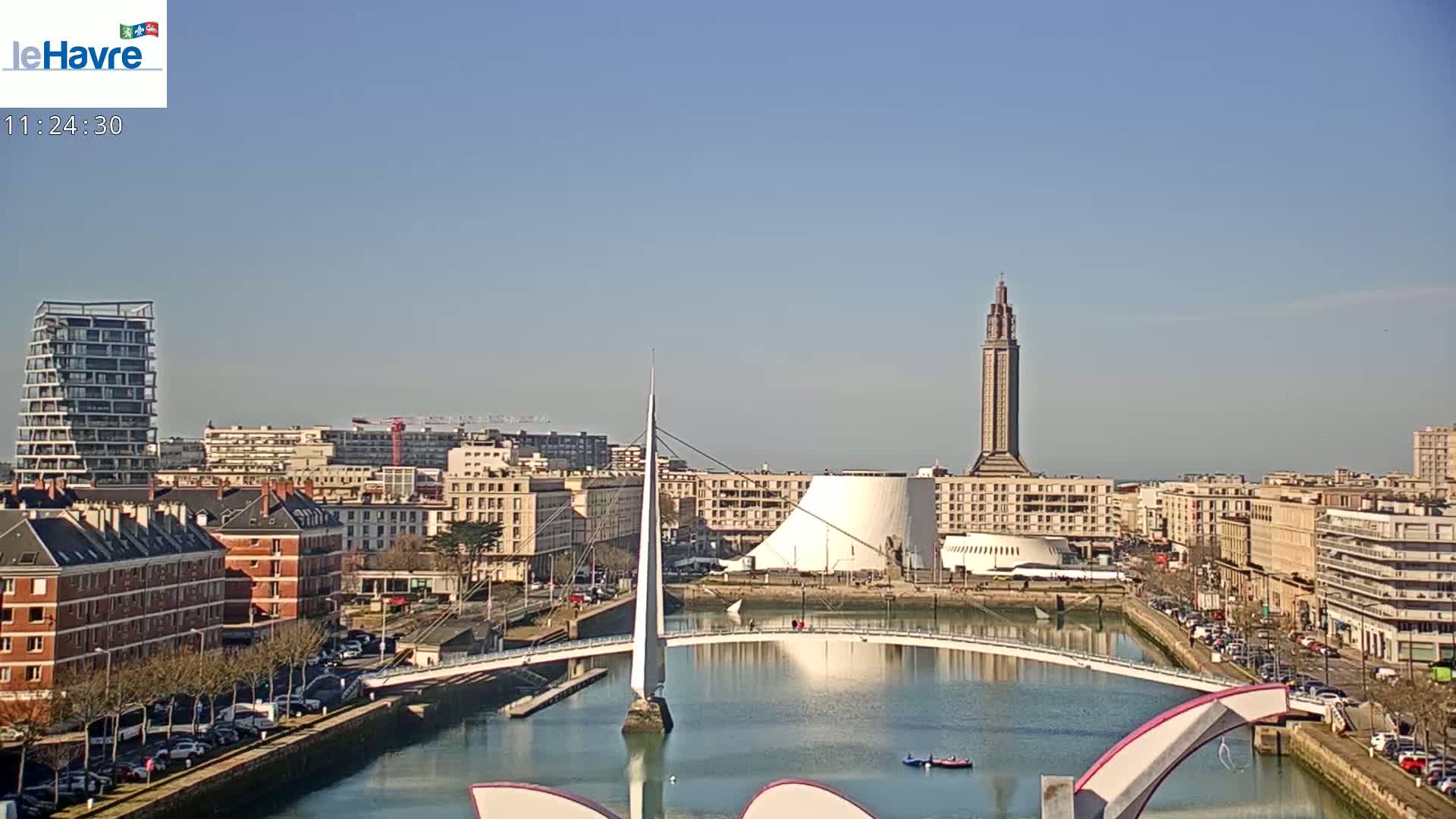 A sunny day reveals a cityscape with a slender bridge spanning a calm waterway, flanked by buildings, including a tall tower and a modern structure with a domed roof.