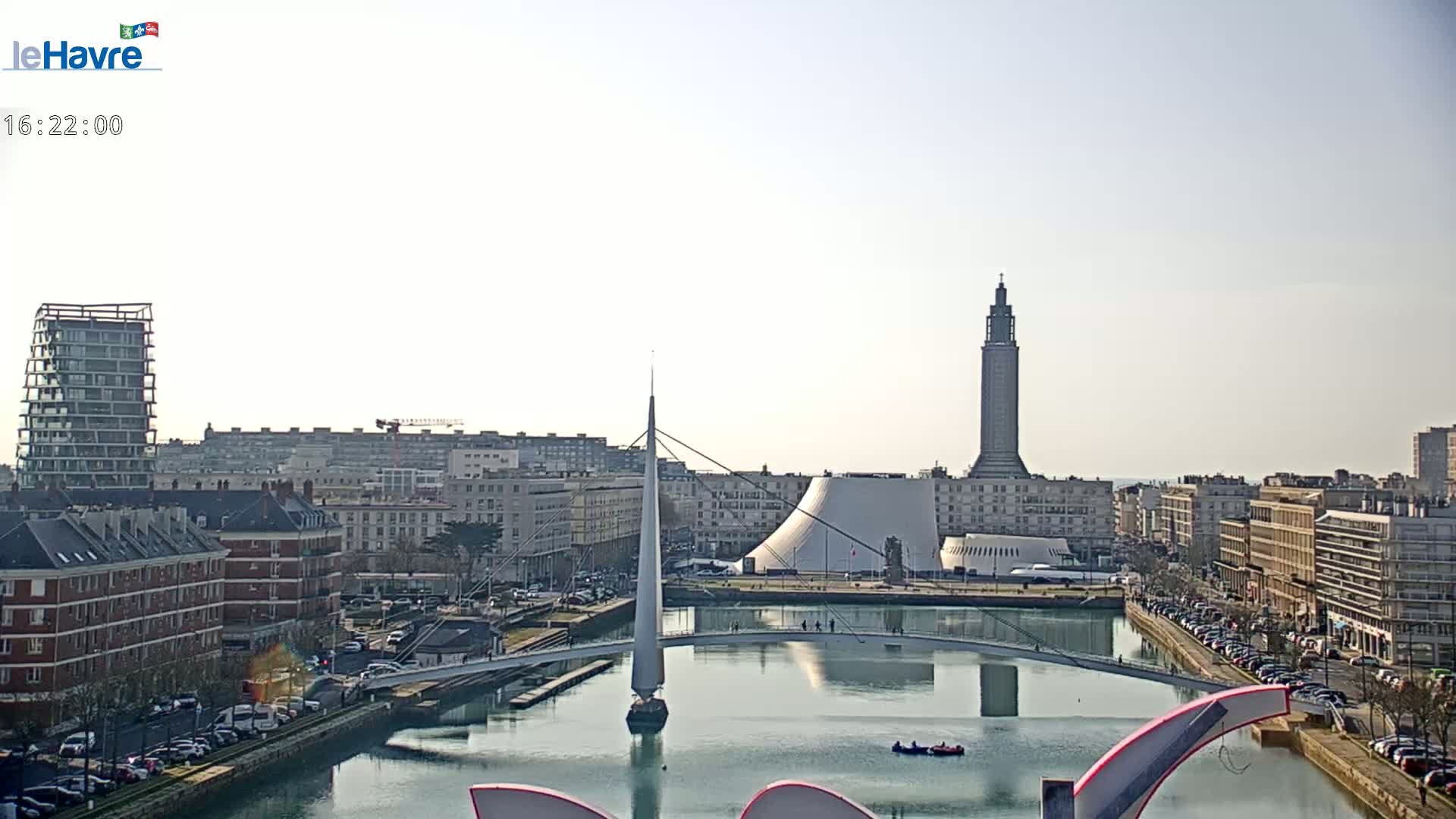 A waterfront city scene on a sunny day shows a canal with a modern pedestrian bridge, buildings, and a tall tower in the background.