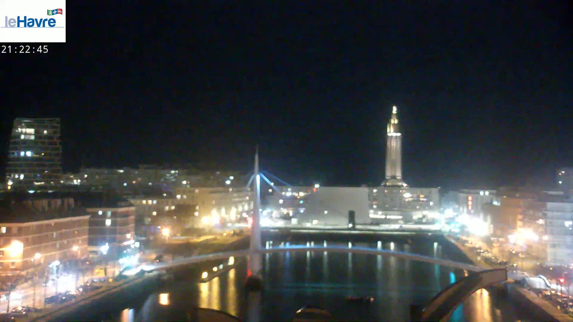 A nighttime view of a city waterfront with a bridge, illuminated buildings, and a tall tower in the background under a clear, dark sky.