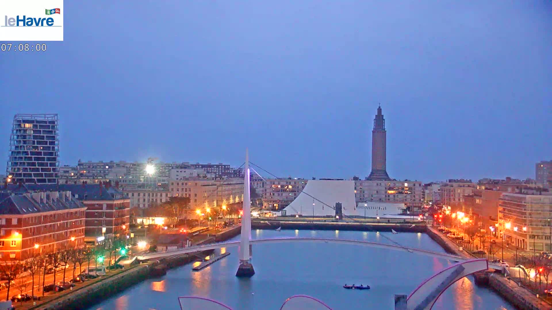 A city waterfront at dusk, featuring a tall tower, a bridge, and calm water.