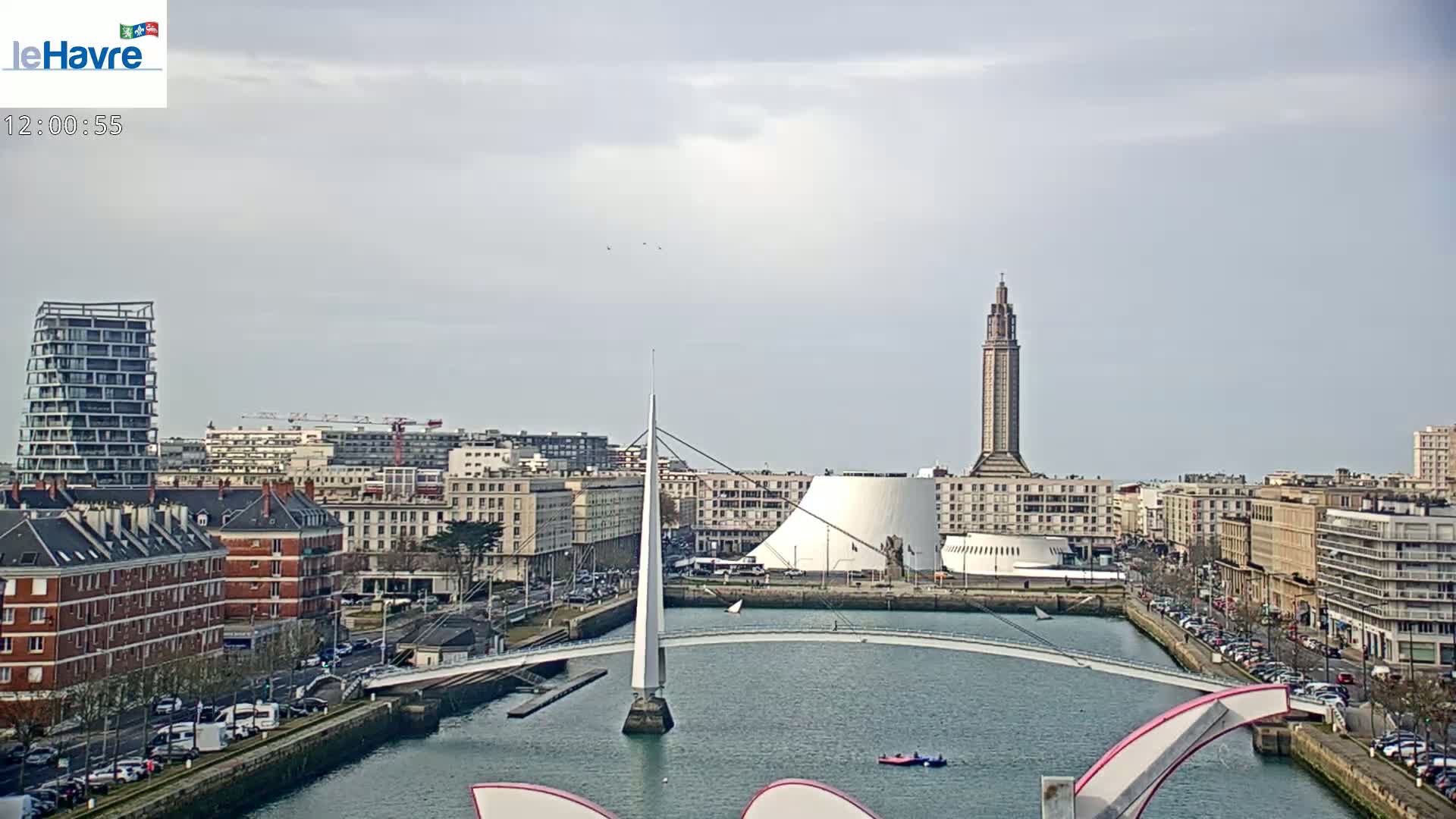 A cityscape with a tall tower and modern buildings overlooks a waterway spanned by a modern white bridge under a cloudy sky.
