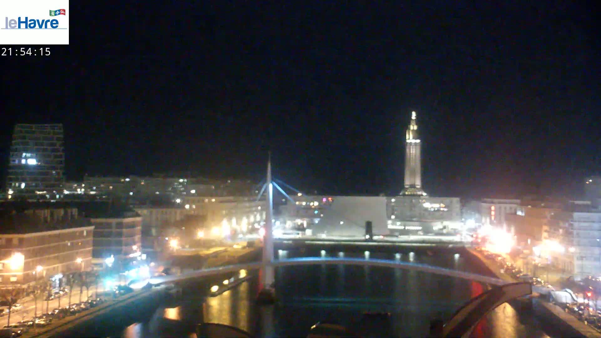 A nighttime view of a city with a bridge crossing a waterway, illuminated buildings, and a tall tower in the background under a clear, dark sky.