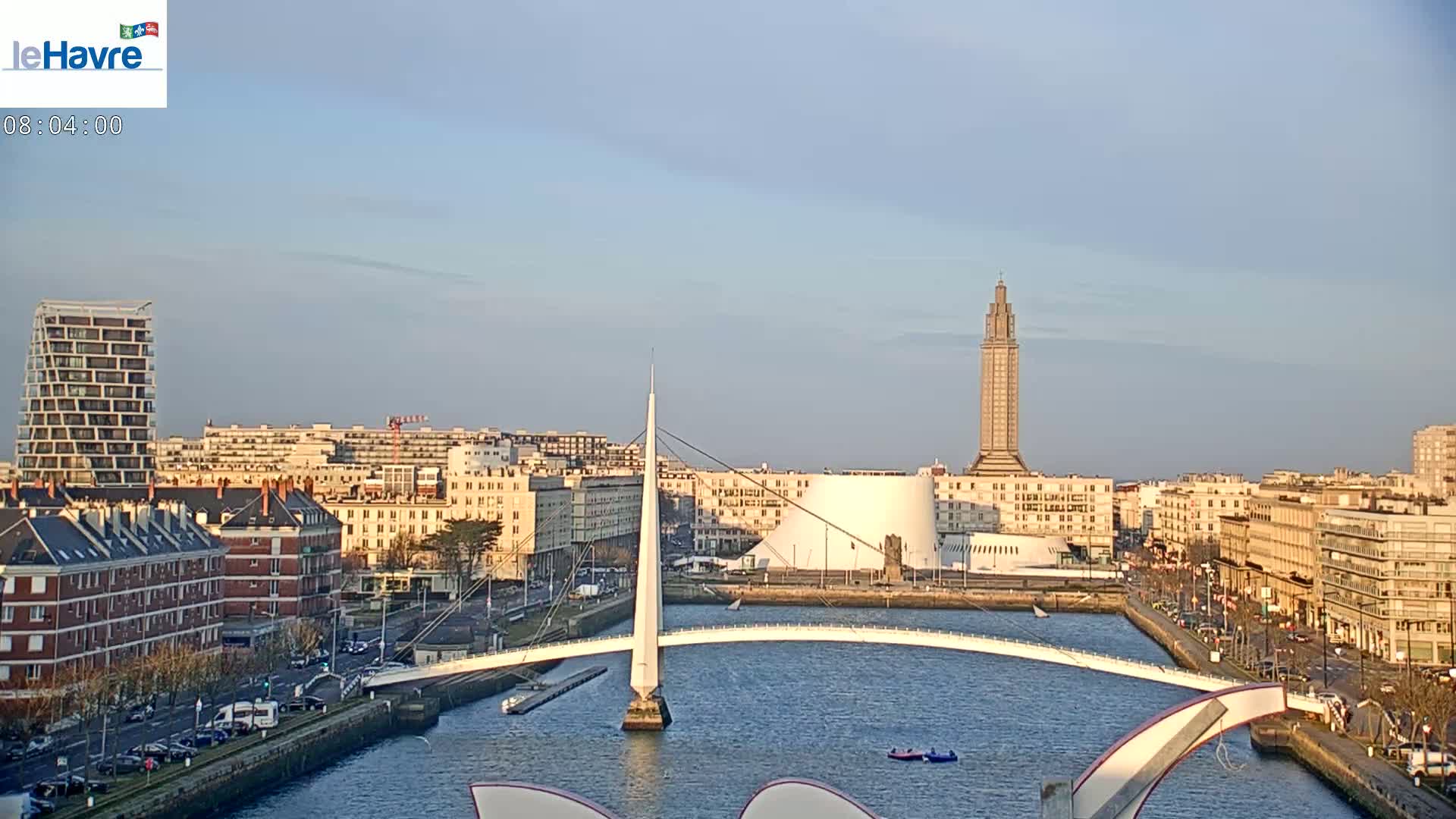 A city skyline with a modern white bridge over a waterway, under a clear, light blue sky.
