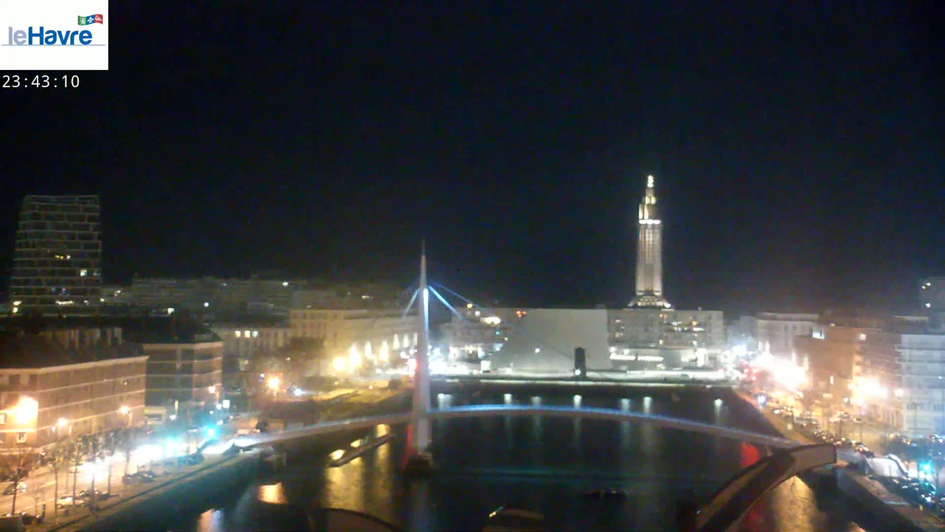 A nighttime cityscape shows a river reflecting lights from buildings and a bridge, under a clear, dark sky.