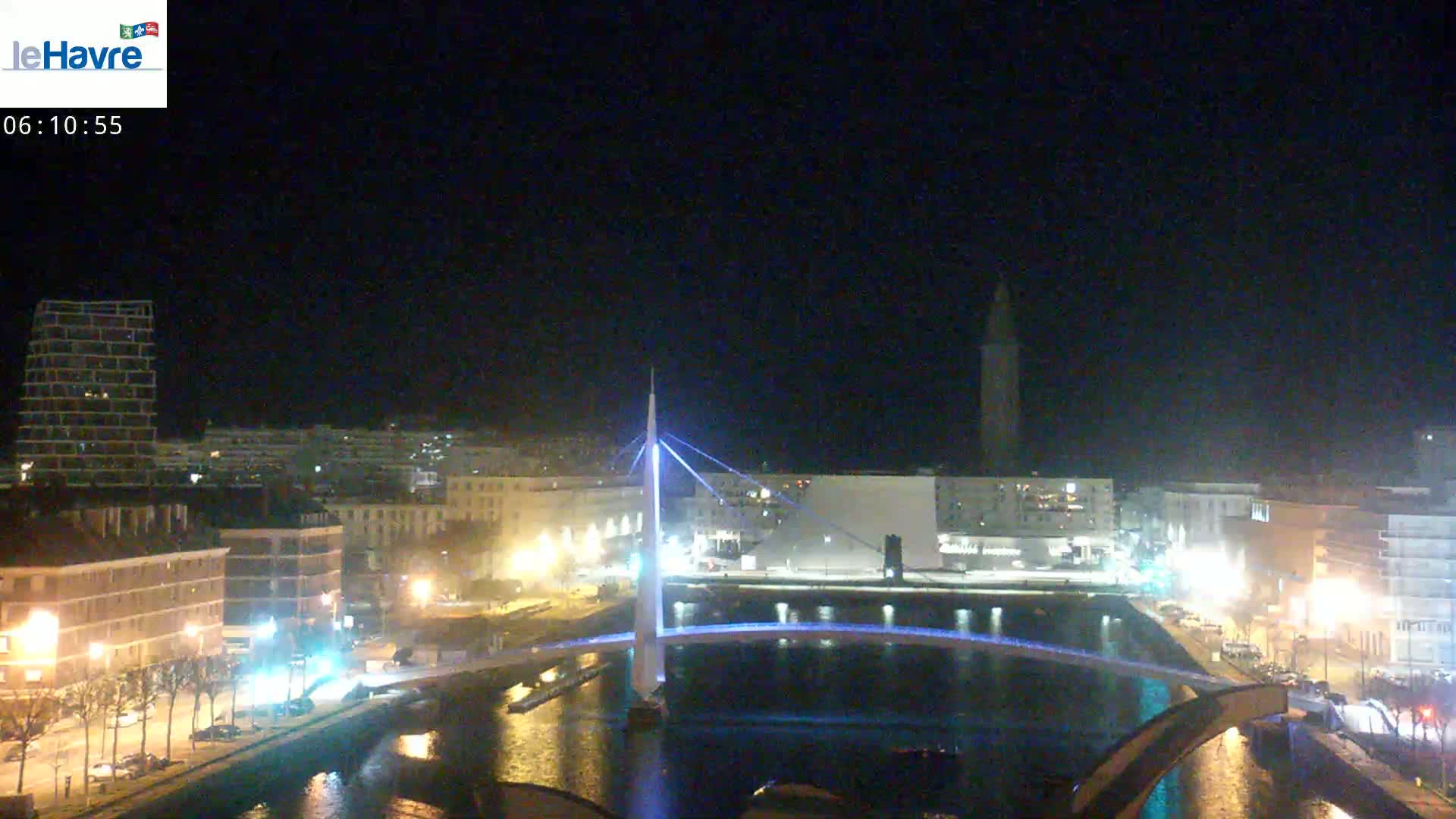 A nighttime view of a city with a lit-up bridge spanning a waterway, buildings lining the water's edge, and a tall structure in the distance under a clear, dark sky.