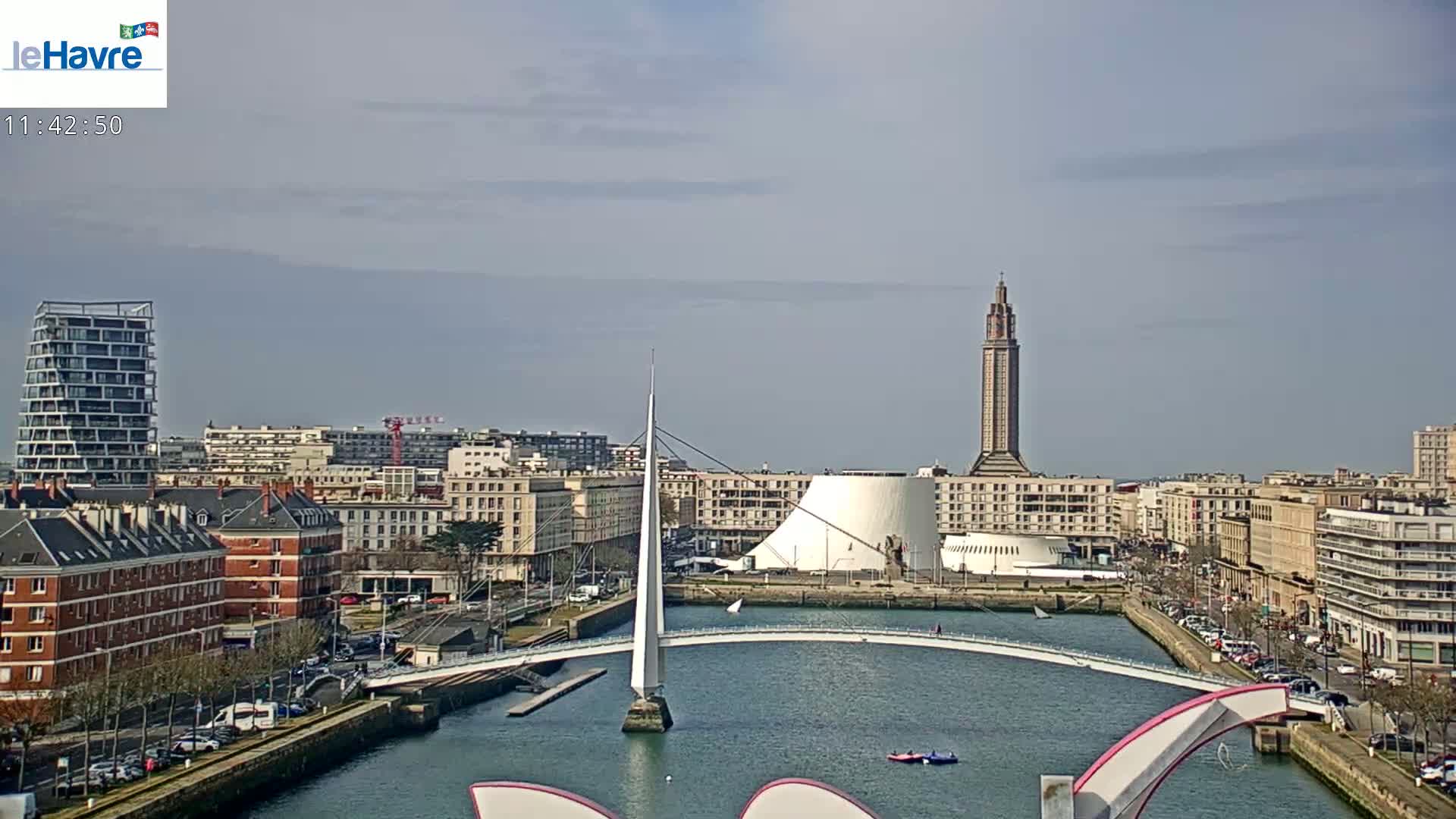 A city waterfront on a partly cloudy day features a modern white pedestrian bridge over a waterway, with a tall tower and other buildings in the background.
