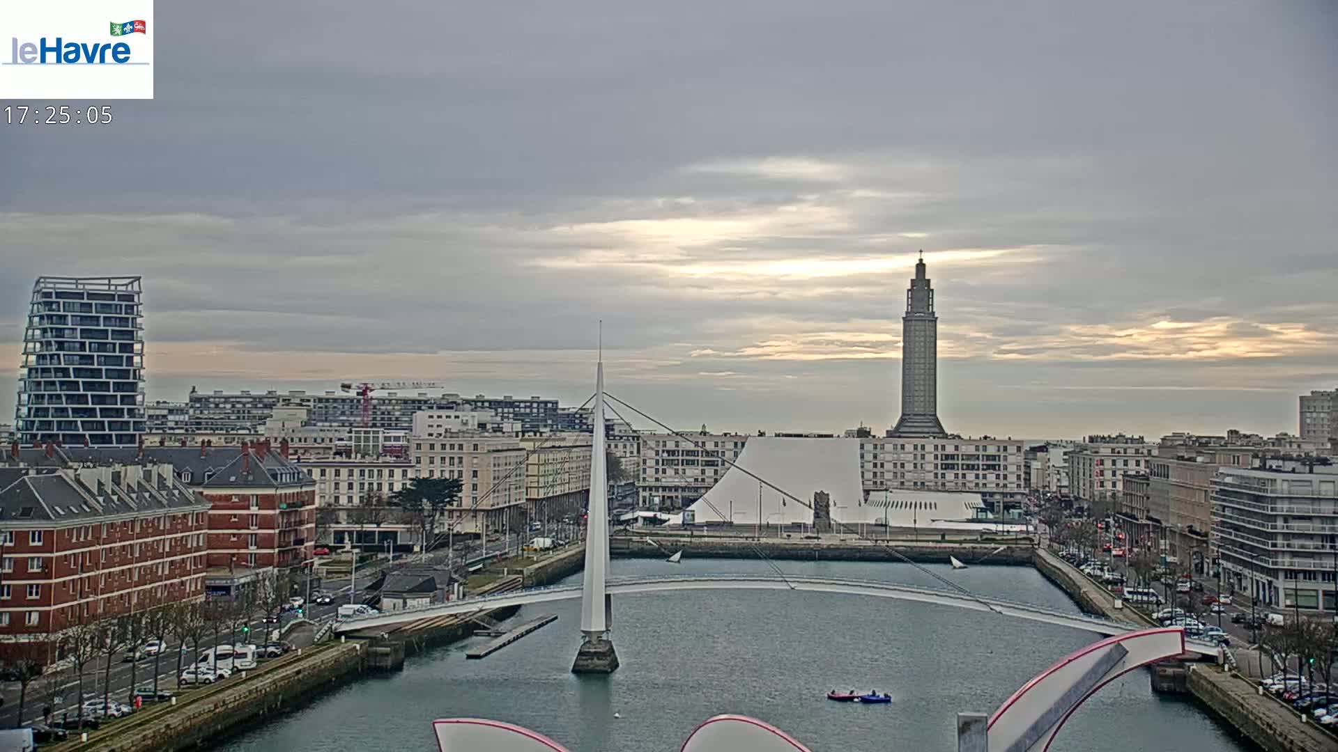 A city skyline with a tall tower and modern buildings is viewed from across a waterway under a partly cloudy sky.