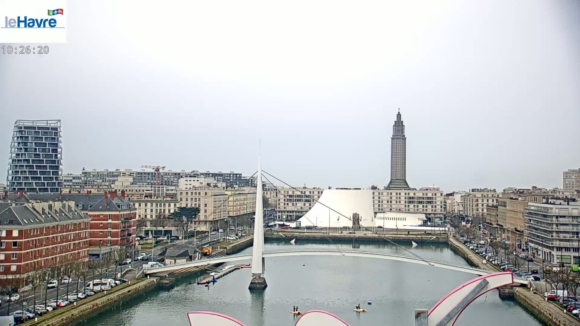 A city waterfront scene on an overcast day shows a canal with a modern white bridge, several buildings, and a tall tower in the distance.