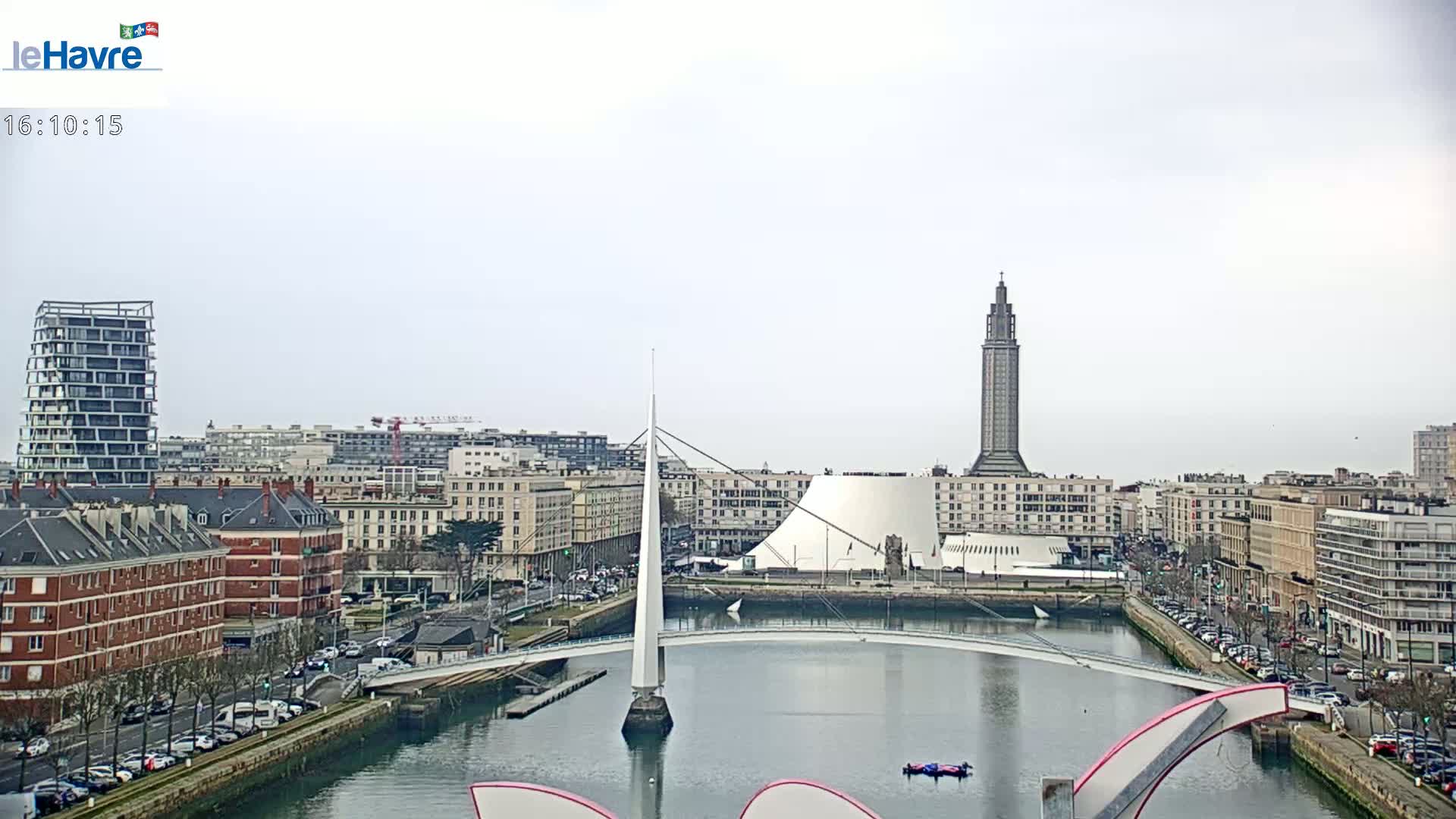 A city waterfront scene on an overcast day shows a bridge, several buildings, and a tall tower, with cars parked along the water's edge.