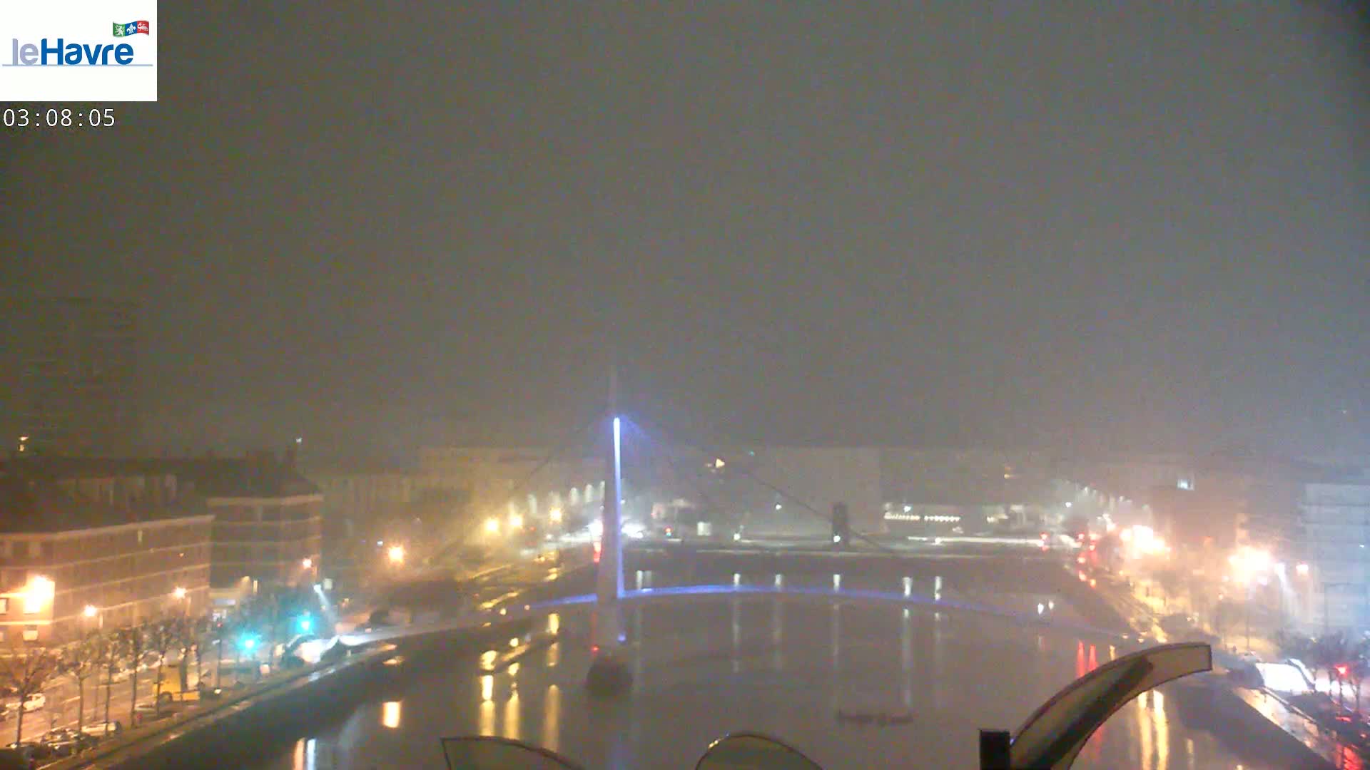 A nighttime view of a city canal with a bridge illuminated in blue under hazy weather conditions.