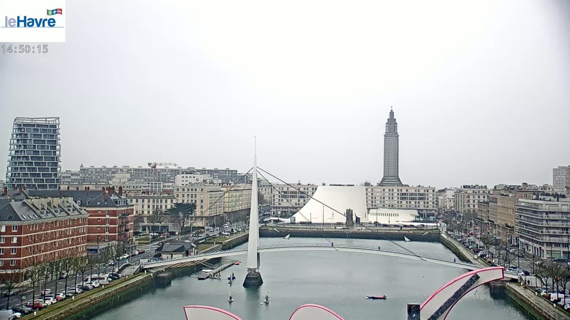 A city waterfront on an overcast day shows a canal with a modern white pedestrian bridge, several buildings, and a tall tower in the background.