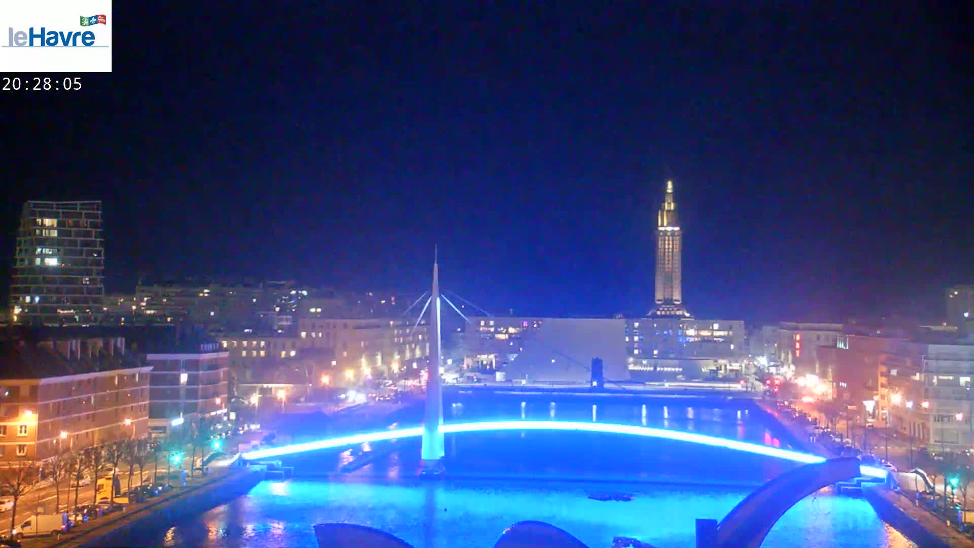 A nighttime cityscape shows a canal with a bridge illuminated in blue, buildings lining the waterfront, and a tall tower in the distance under a clear, dark sky.