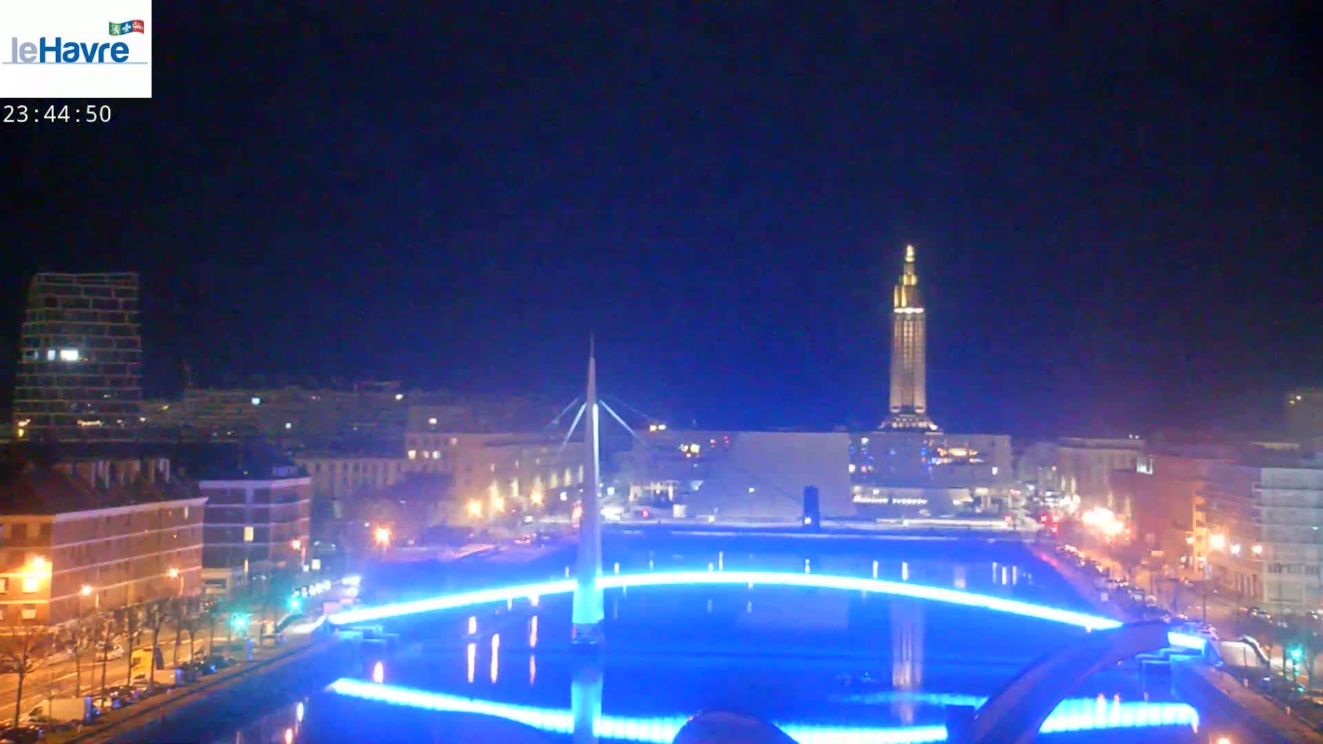 A nighttime cityscape with a tall, illuminated tower in the background, a bridge with blue lights illuminating a reflecting pool in the foreground, and clear skies.