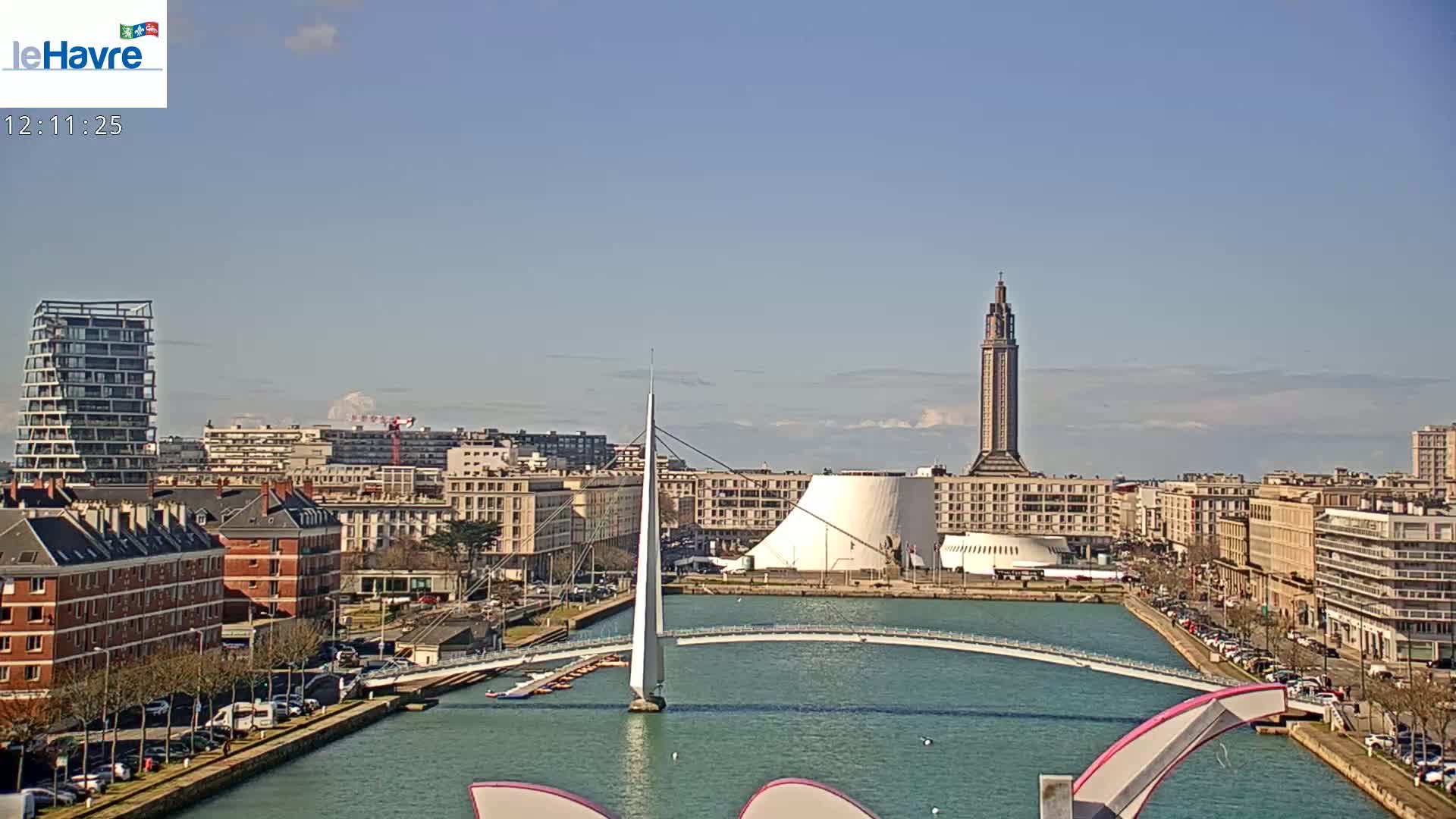 A city waterfront scene on a sunny day shows a slender white pedestrian bridge spanning a canal, with buildings, a tall tower, and a modern white building in the background.