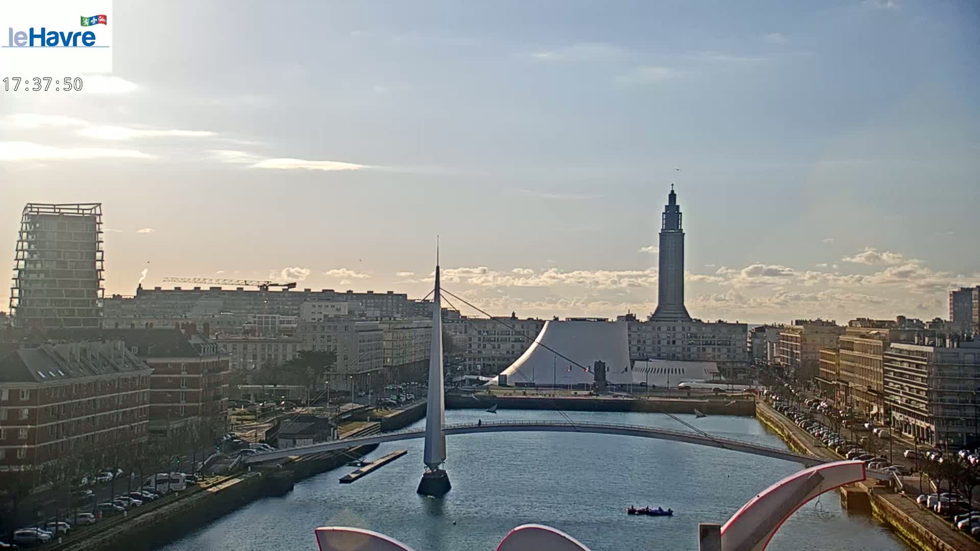 A partly sunny day shows a cityscape with a canal, several buildings, a tall tower, and a modern footbridge.