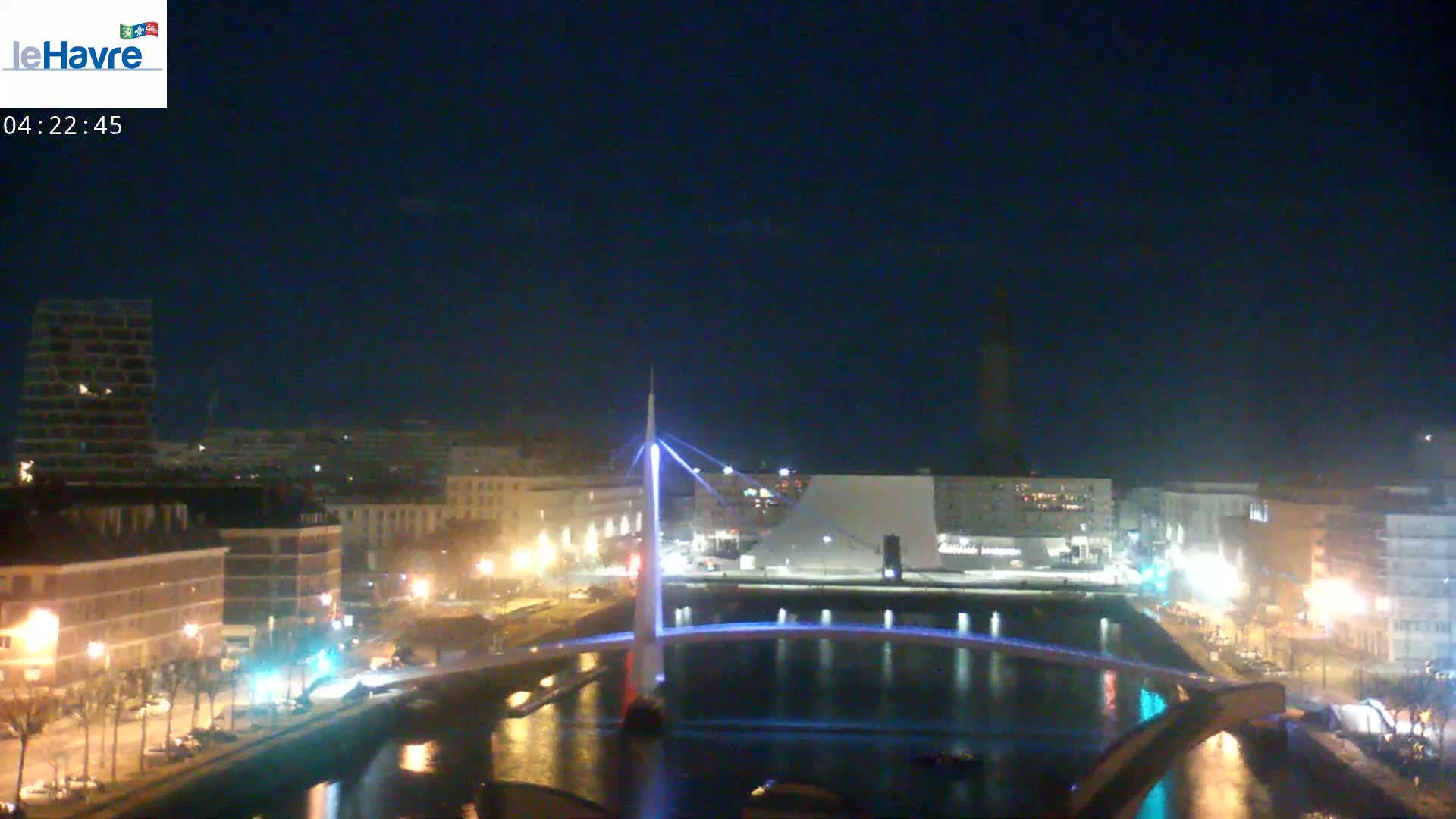 A nighttime view of a city waterfront with a modern, illuminated bridge spanning a calm waterway, under a clear, dark sky.