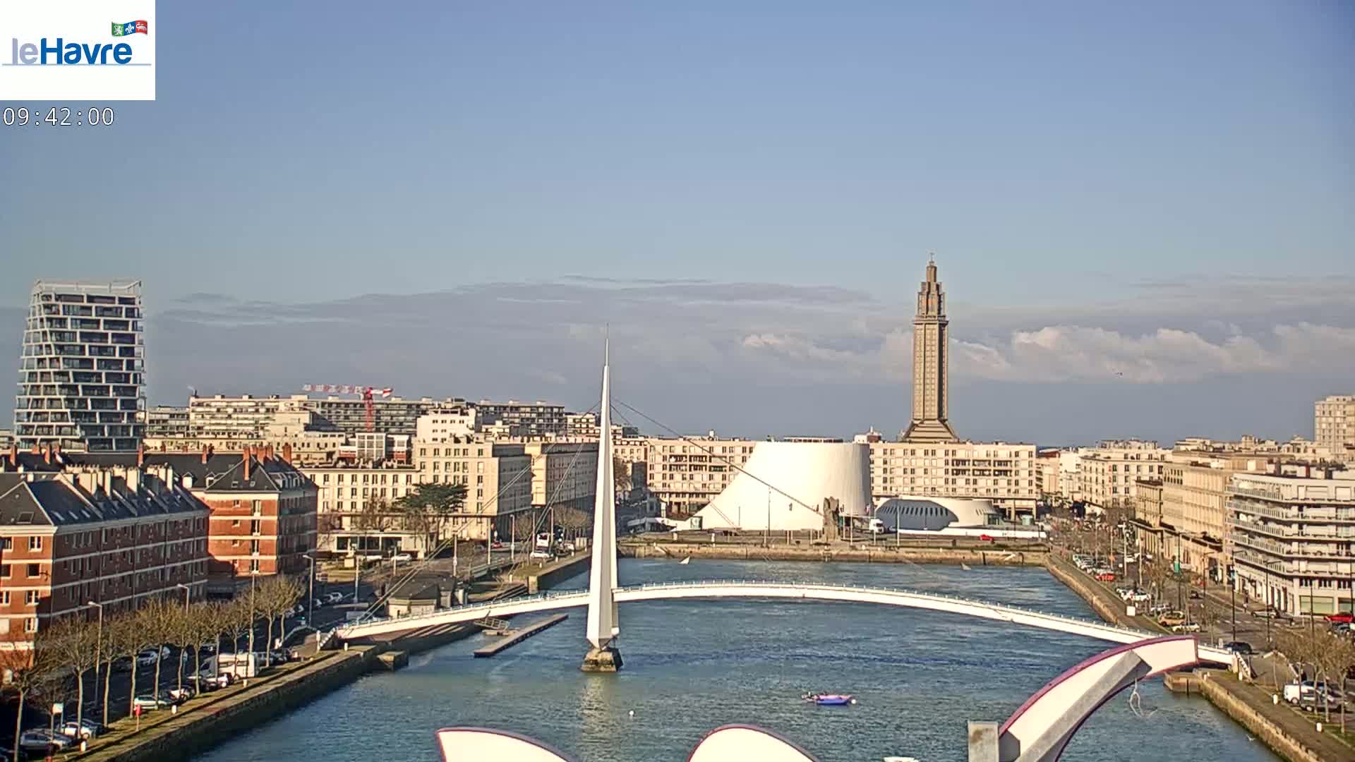 A city waterfront scene on a partly cloudy day shows a modern bridge arching over a waterway, with buildings, a tall tower, and a curved white structure in the background.