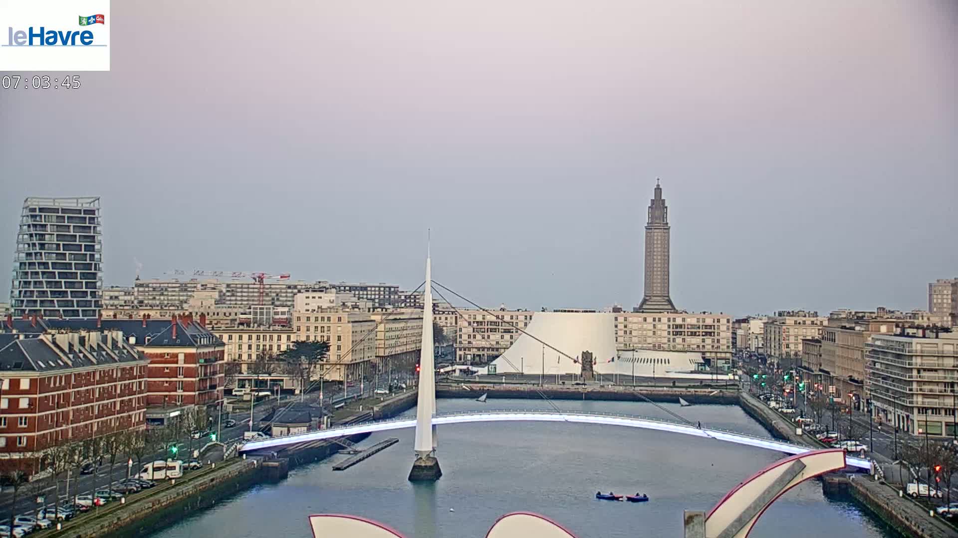 A city skyline with a modern white bridge over a waterway, under a pale, overcast sky.