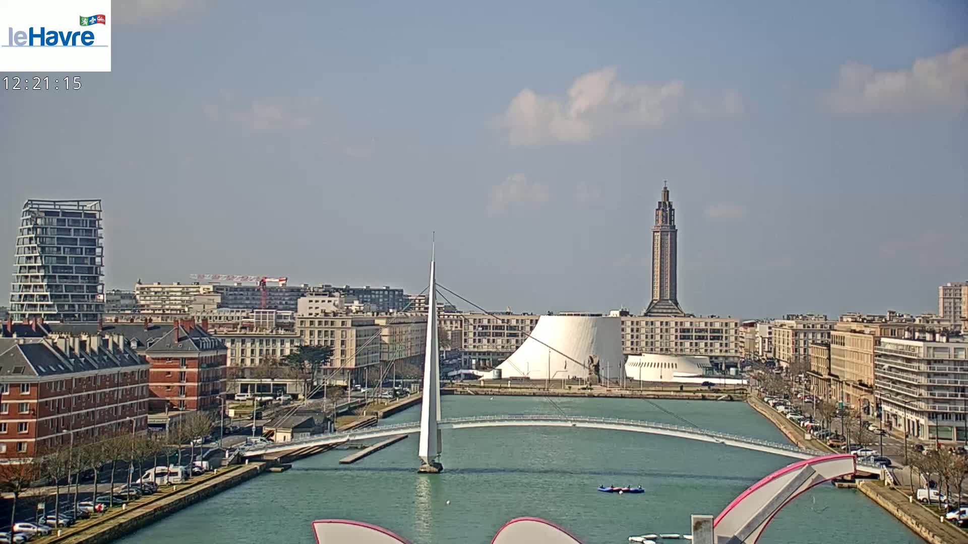 A partly cloudy day reveals a city waterfront with a modern bridge, a tall tower, and several buildings lining a canal.