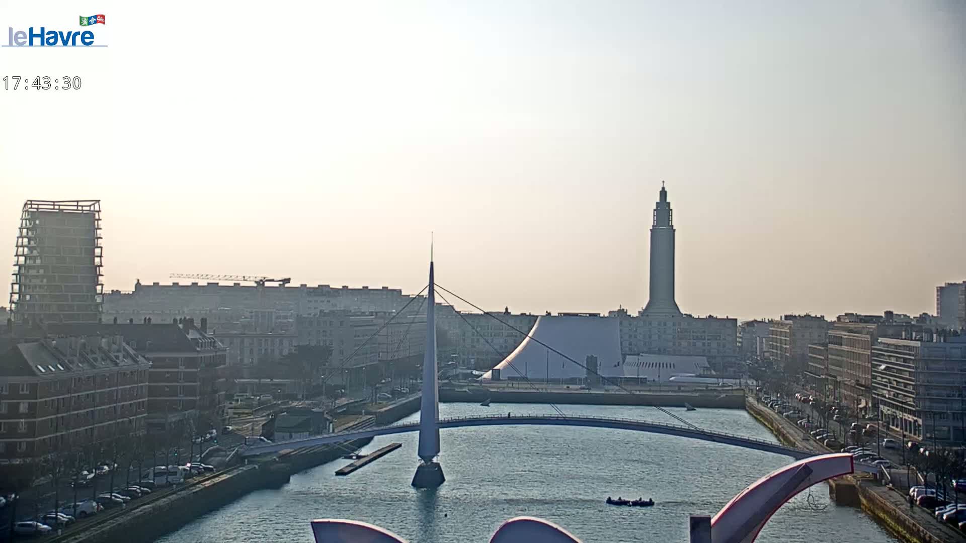 A hazy, sunny cityscape features a canal with a modern bridge, several buildings, and a tall tower in the background.