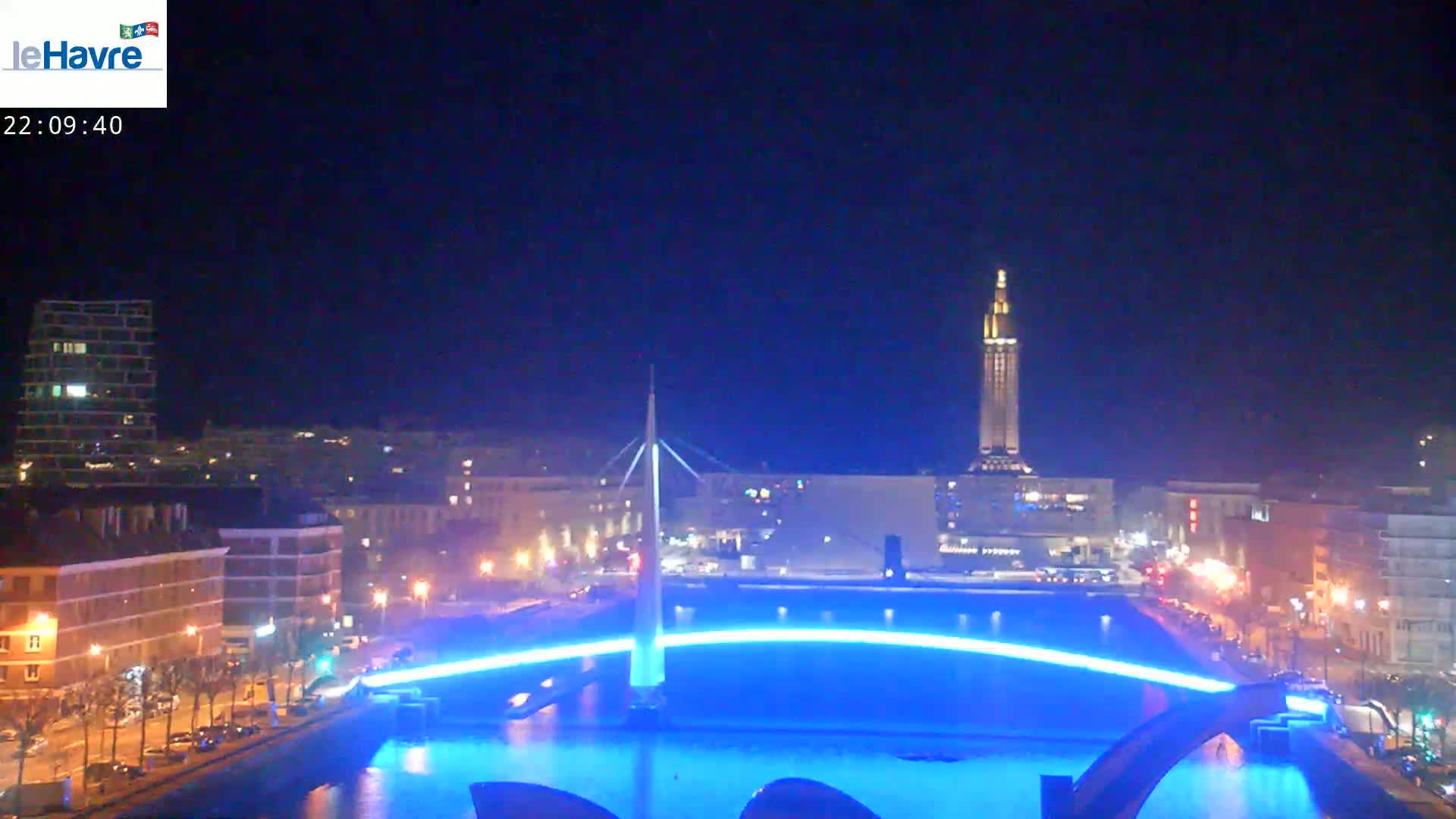 A nighttime cityscape with a bridge illuminated in bright blue, reflecting in the water below, and a tall tower visible in the background under a clear, dark sky.