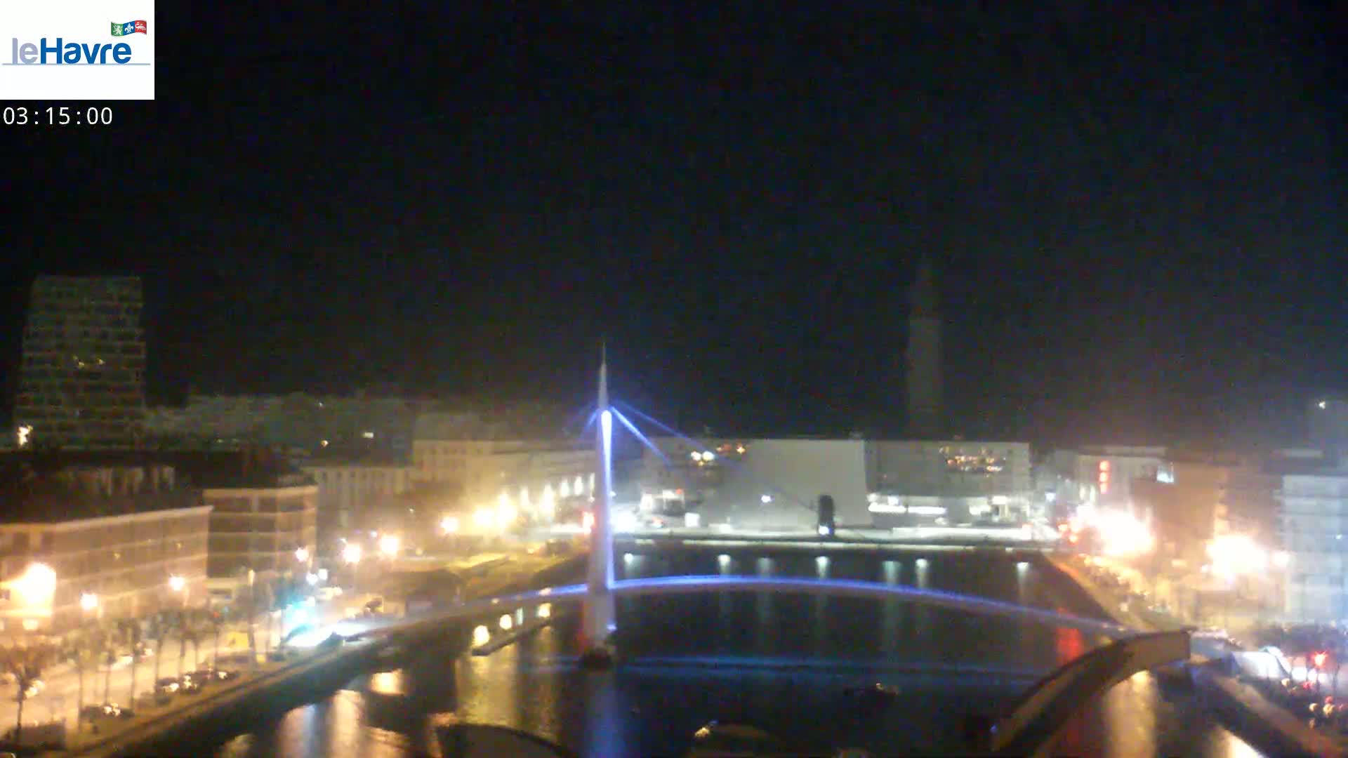 A nighttime view of a city canal with a modern illuminated bridge, buildings, and streetlights along the waterfront under a clear, dark sky.