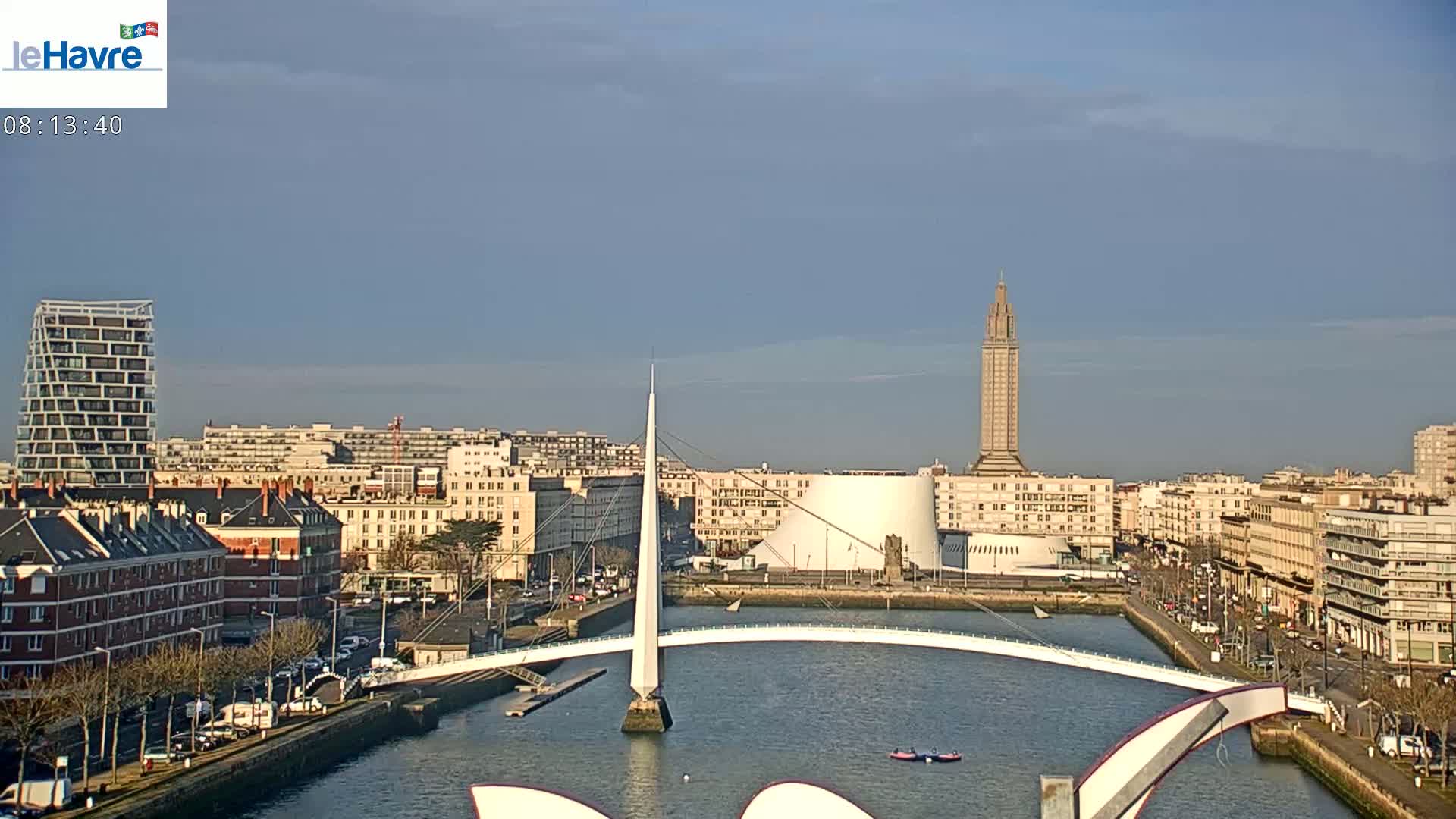 A city waterfront scene on a partly sunny day shows a white pedestrian bridge arching over a calm waterway, with buildings, a tall tower, and a modern structure along the shore.