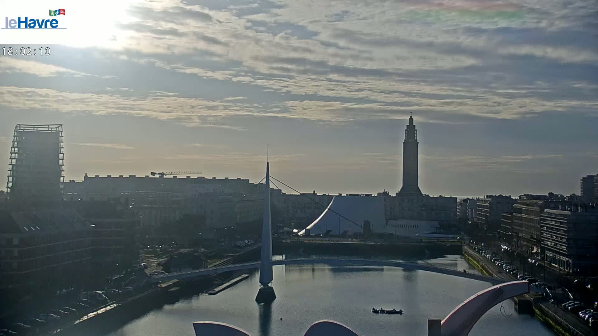 A partly cloudy sunrise illuminates a cityscape with a tall tower, a modern building, and a canal, showcasing a bridge and docked boats.