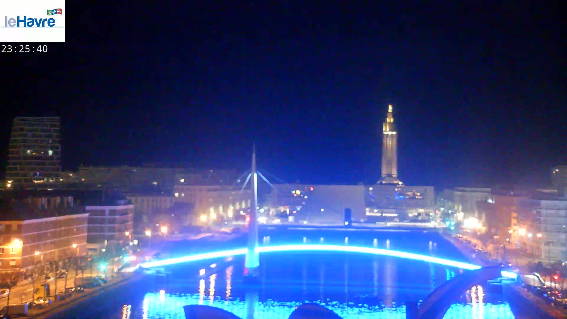 A nighttime cityscape with a body of water illuminated in blue, featuring a bridge and a tall tower in the background.