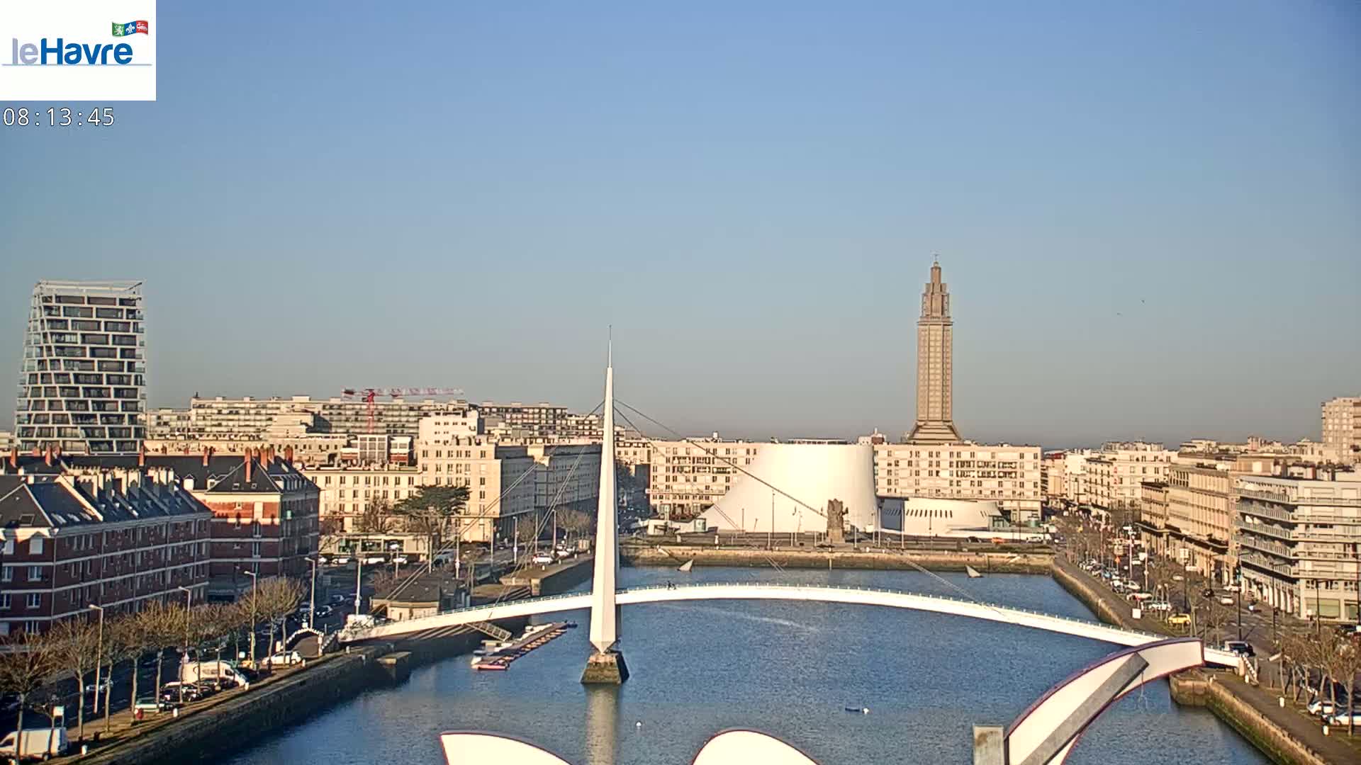 A cityscape with a modern white bridge over a waterway, under a clear blue sky, features a tall tower and various buildings.