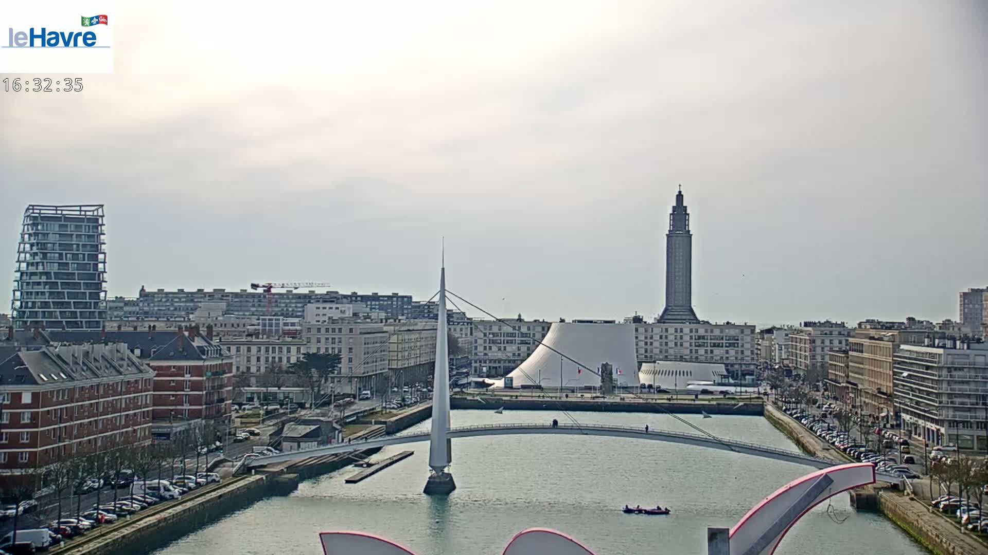 A city waterfront scene under a cloudy sky shows a canal with a pedestrian bridge, buildings, and a tall tower in the distance.