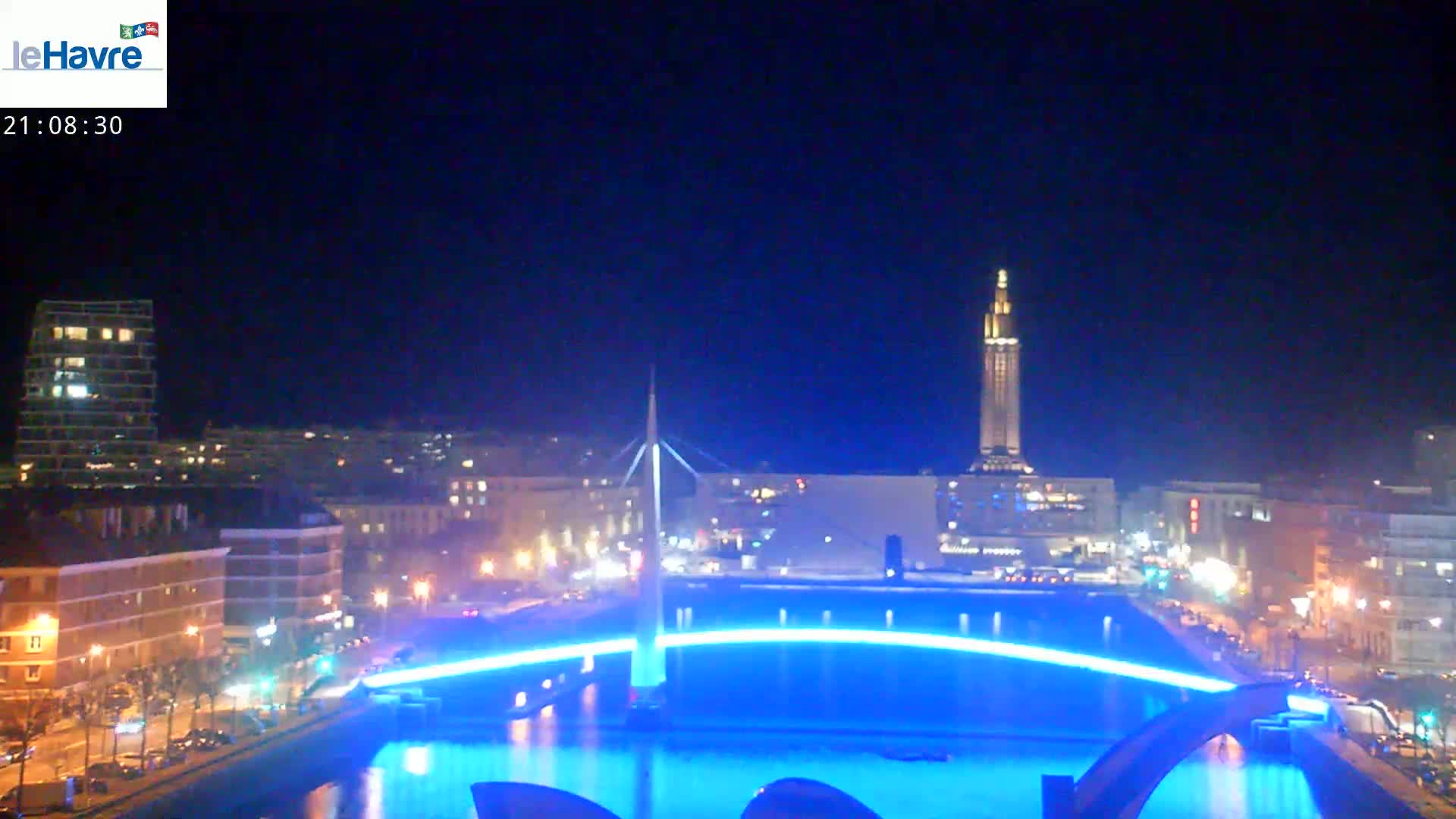 A nighttime cityscape with a bridge illuminated in blue light, reflecting in a body of water, and a tall tower in the background under a clear, dark sky.