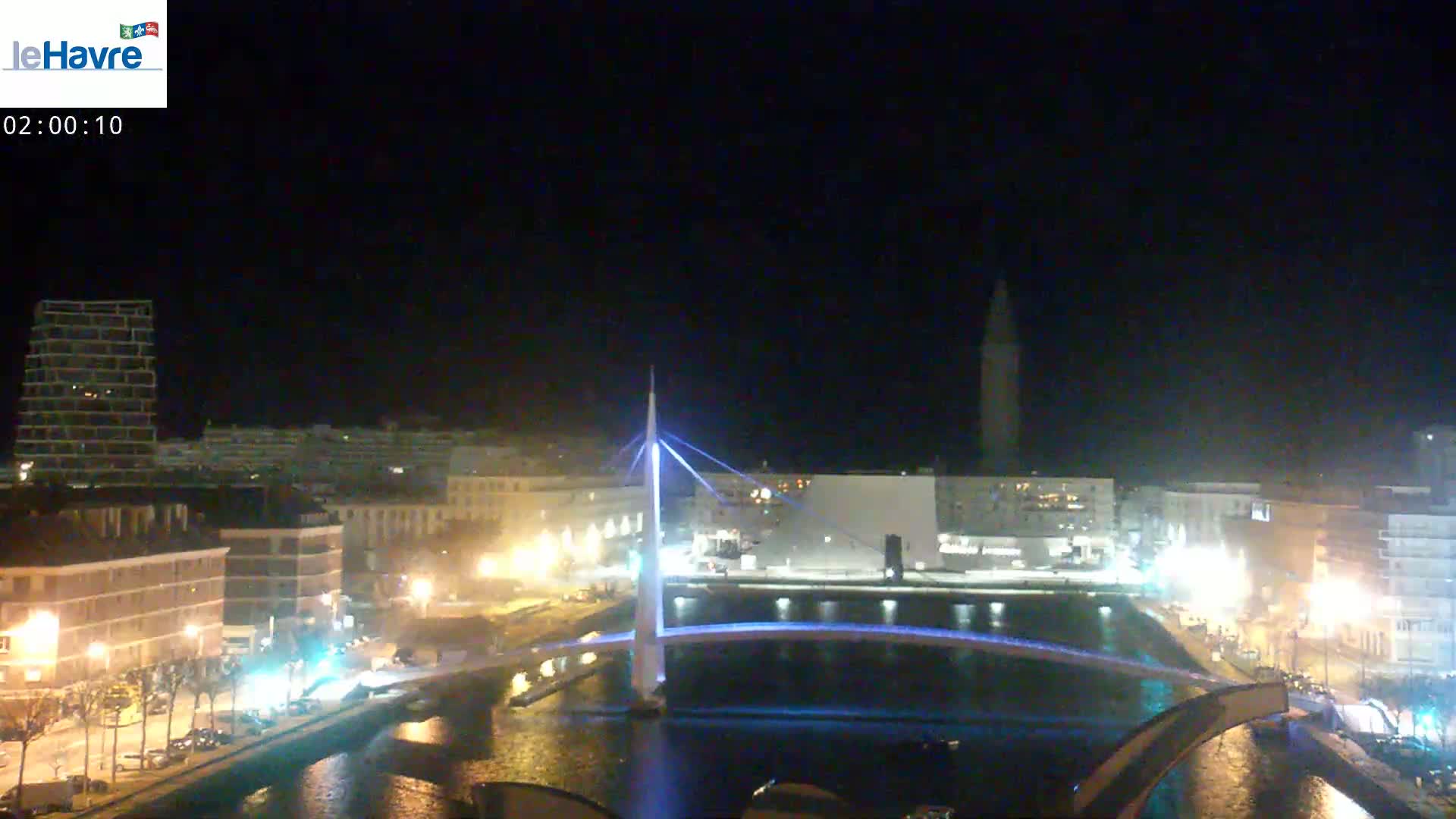 A nighttime view of a city's waterfront, featuring a modern bridge illuminated with blue lights, canals, and buildings along the shoreline under a clear, dark sky.