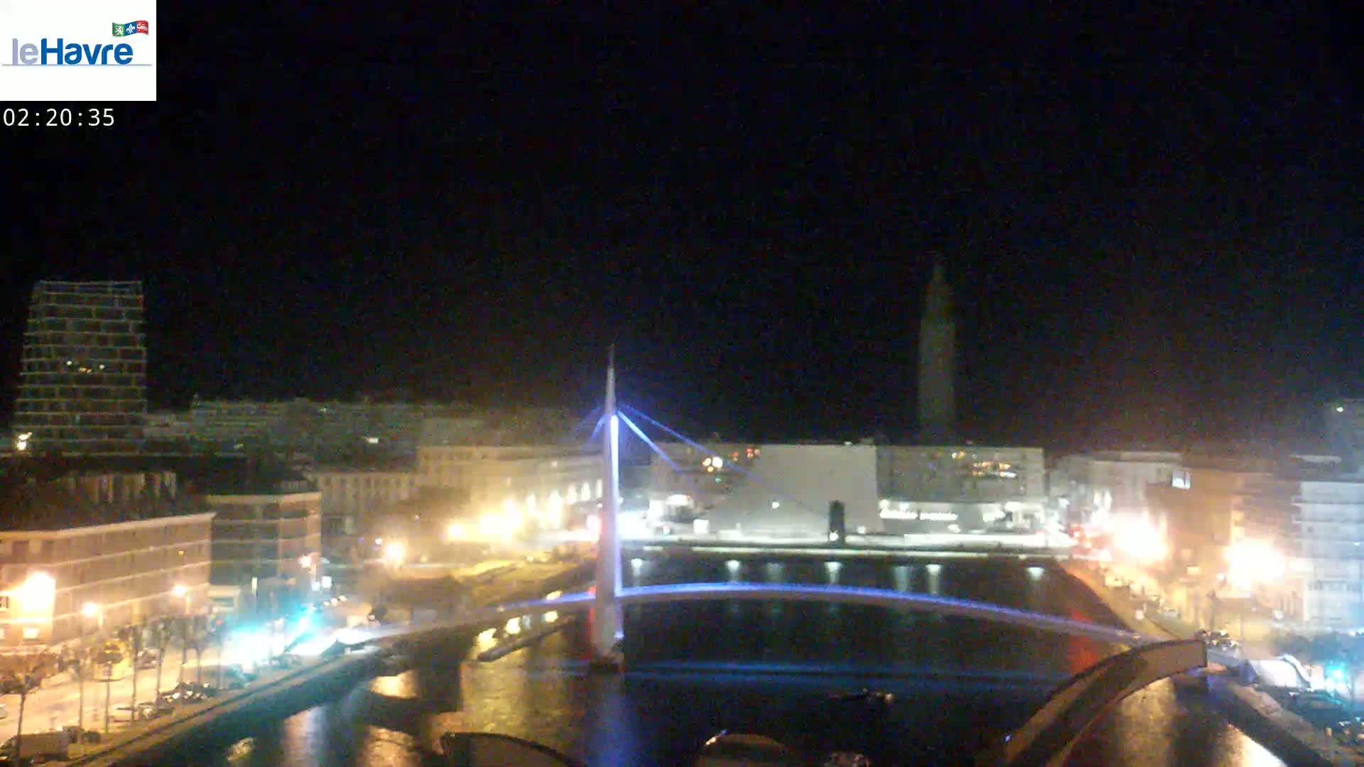 A nighttime view of a city waterfront, featuring a lit-up bridge spanning a canal, with buildings and streetlights illuminating the scene under a clear, dark sky.