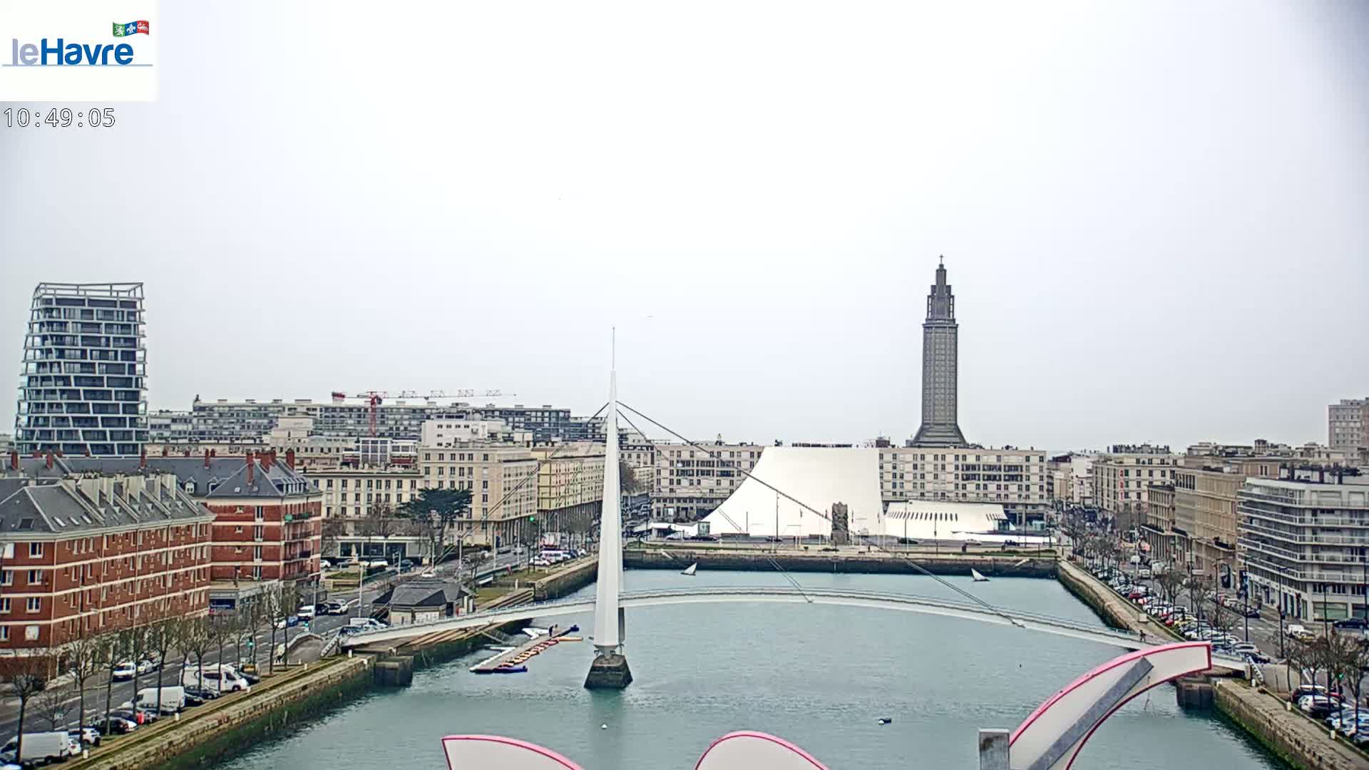 A city waterfront on an overcast day, showing a canal with a modern footbridge, various buildings including a tall tower, and parked cars along the streets.
