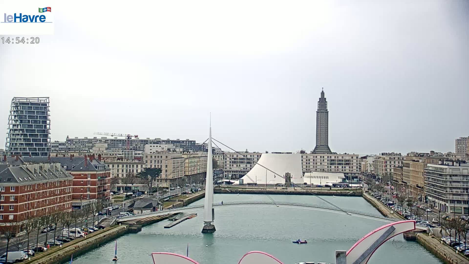 A city waterfront scene under an overcast sky shows a slender bridge spanning a waterway, with buildings and a tall tower in the background.