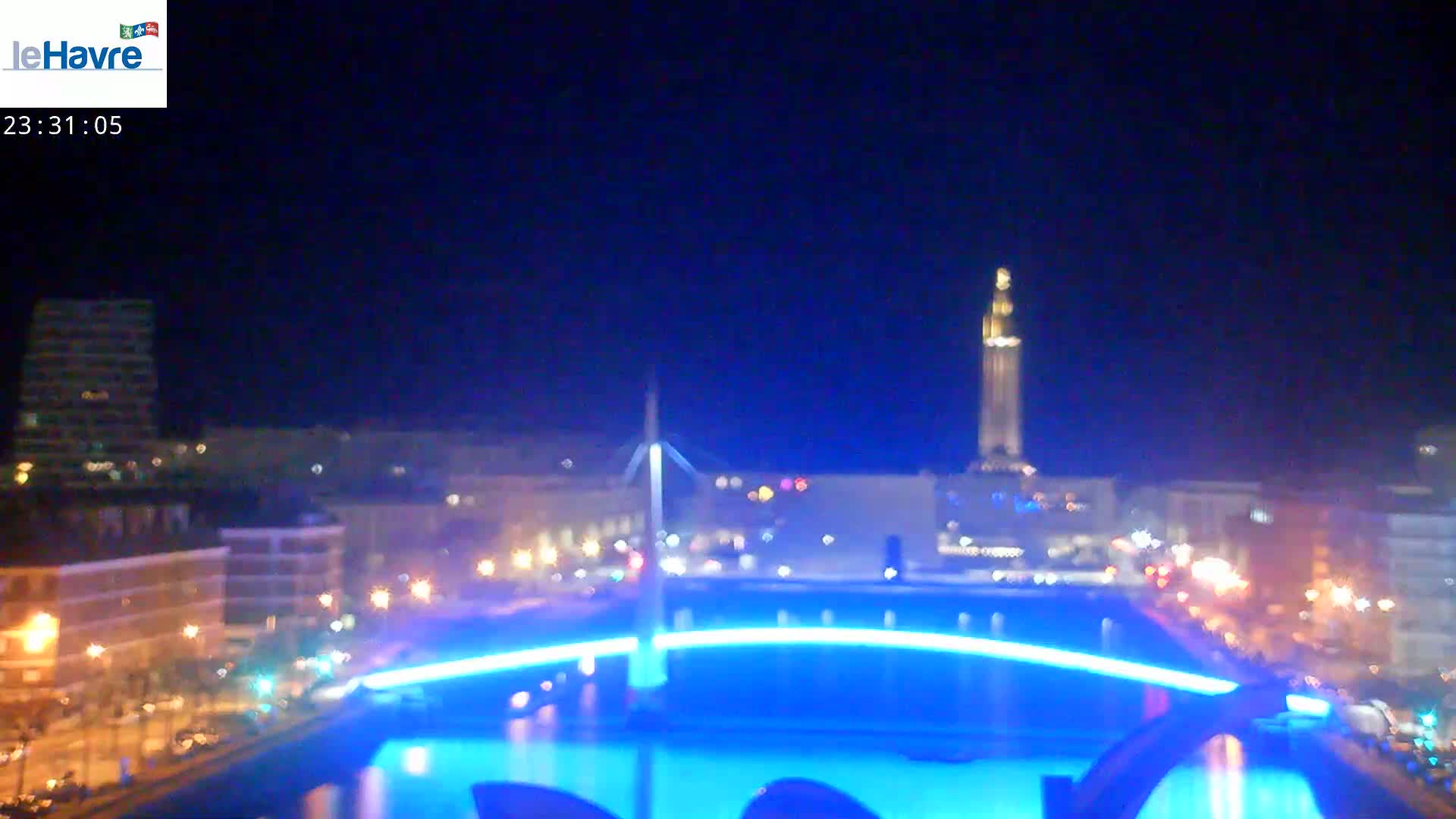 A nighttime city scene shows a brightly lit bridge and fountain in the foreground, with buildings and a tall tower in the background under a dark, clear sky.