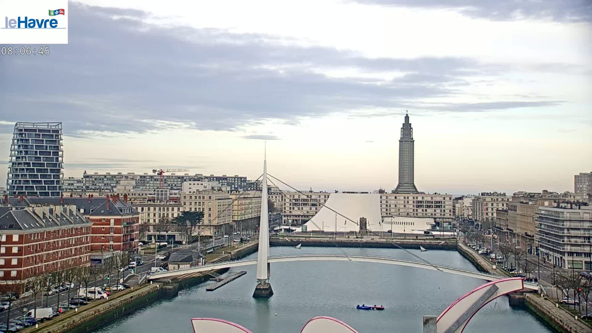 A city skyline with overcast skies, featuring a tall tower, modern buildings, a white pedestrian bridge over a canal, and a small sailboat in the water.