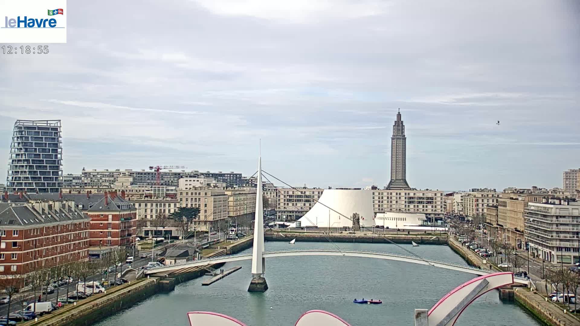A cityscape with a modern bridge over a waterway, under a cloudy sky, features a tall tower and several buildings.