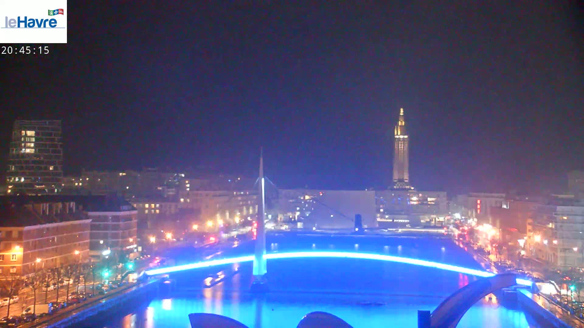 A nighttime cityscape with a brightly lit bridge arching over a body of water, illuminated buildings lining the waterfront, and a tall tower visible in the distance under a clear, dark sky.