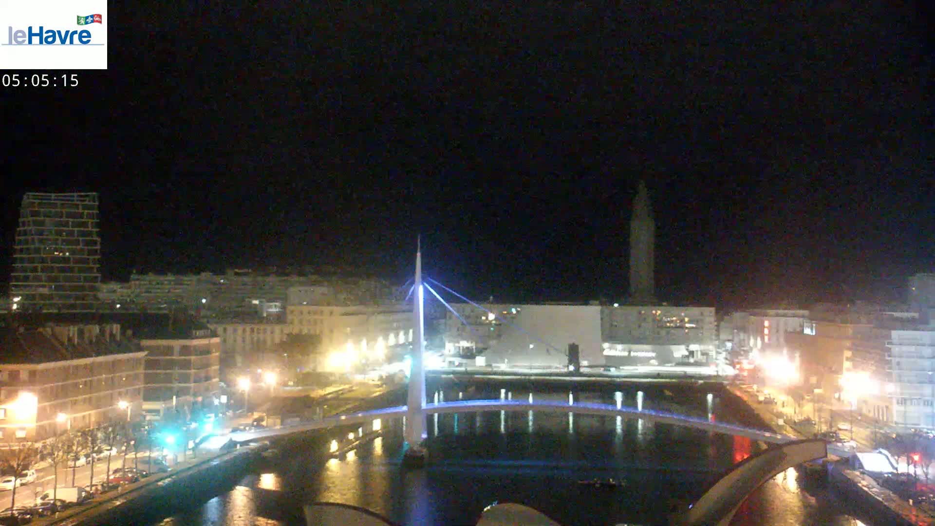 A nighttime view of a city with a bridge illuminated over a calm waterway, buildings lining the waterfront, and a tall structure visible in the distance under a clear, dark sky.