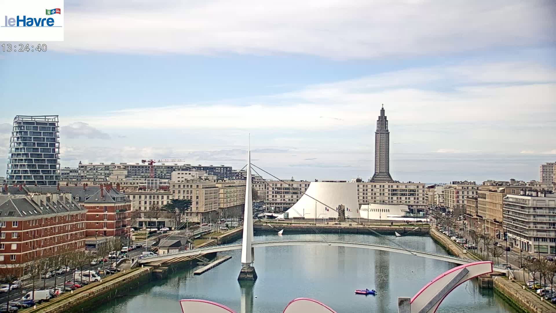 A city harbor scene on a partly cloudy day shows a white modern bridge spanning a waterway, with buildings, a tall tower, and a white domed structure in the background.