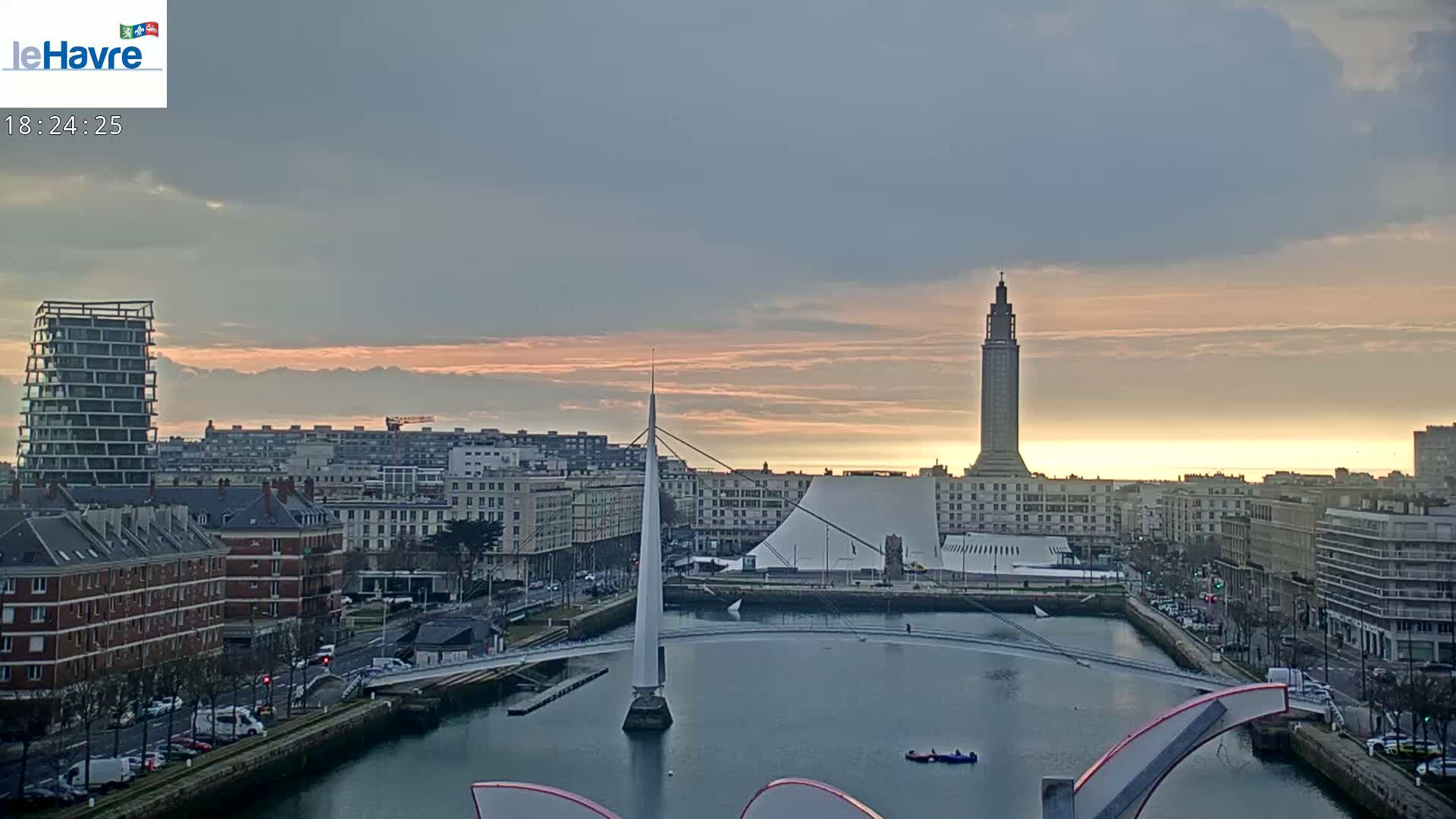 A city skyline at sunset, featuring a tall tower, a modern building with a curved roof, and a slender bridge crossing a waterway, under a mostly cloudy sky.