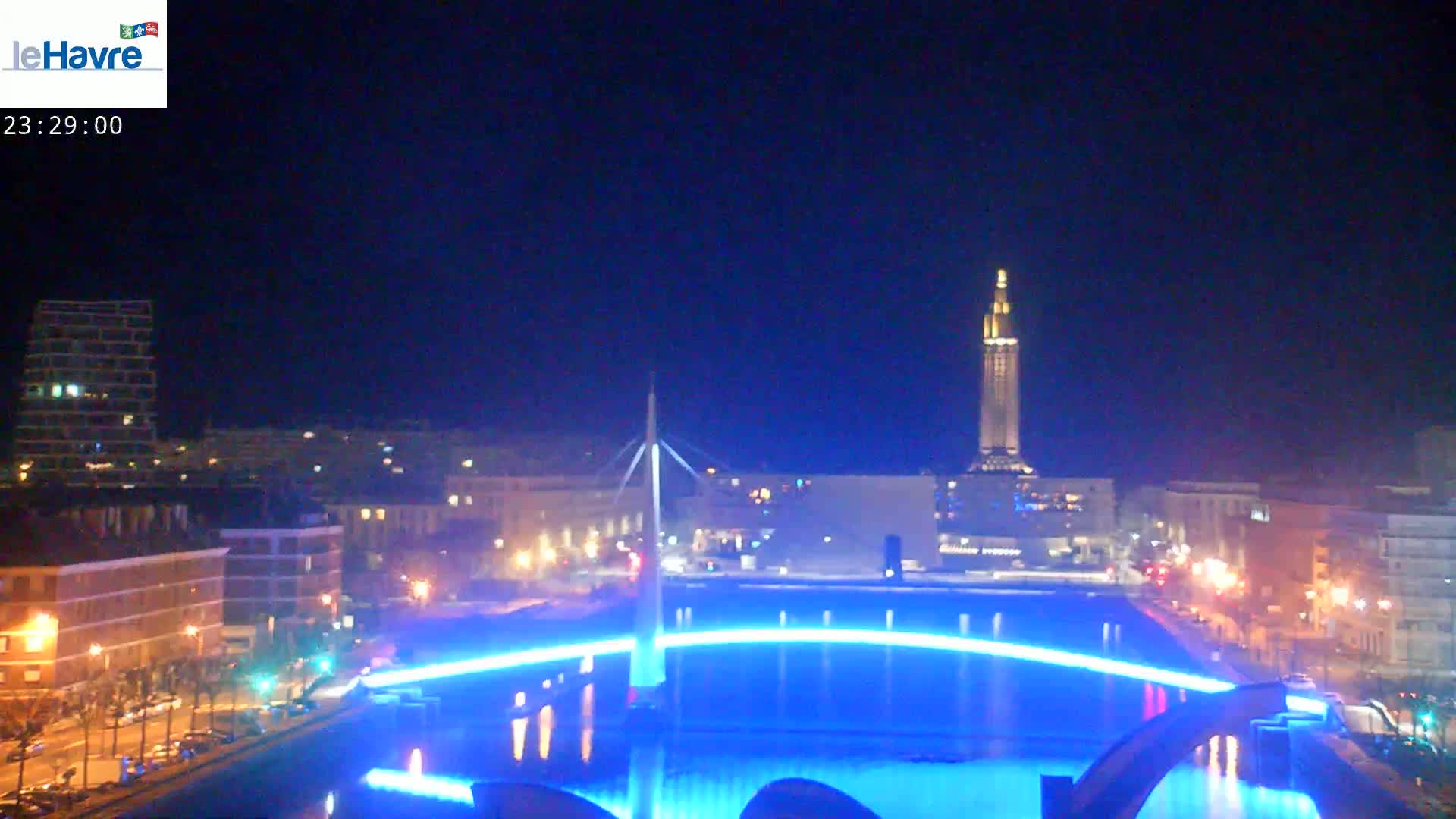 A nighttime cityscape featuring a body of water with blue lights illuminating a bridge and surrounding structures, under a clear sky.