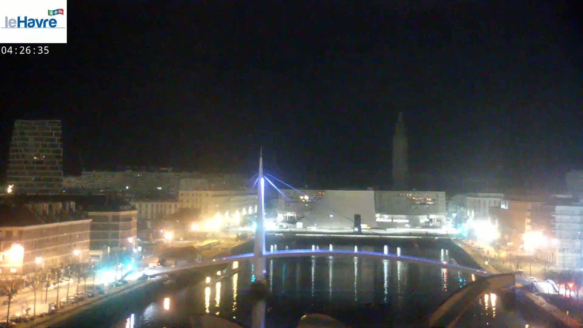 A nighttime view of a city with a lit-up bridge spanning a calm body of water, with buildings and streetlights visible along the waterfront under a clear, dark sky.