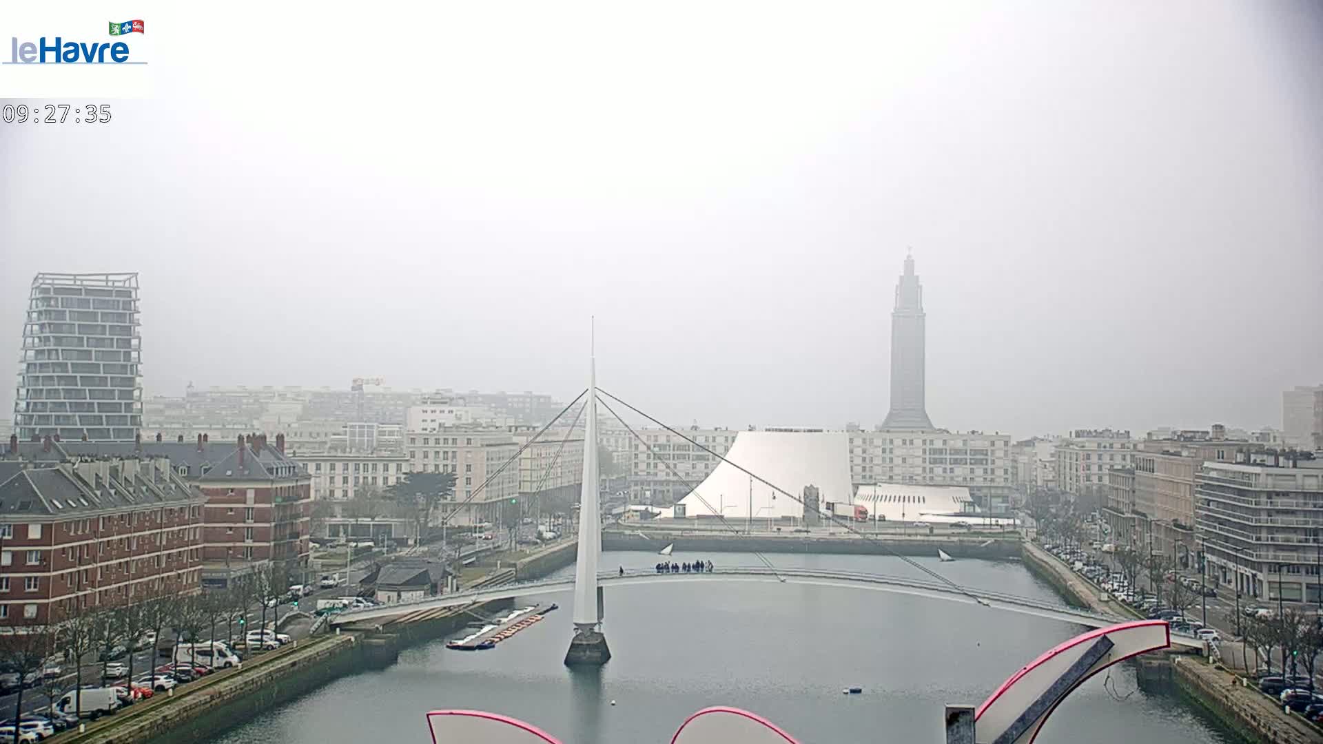 A foggy cityscape shows a canal with a modern pedestrian bridge and several buildings, including a tall tower and a contemporary structure with a rounded roof.