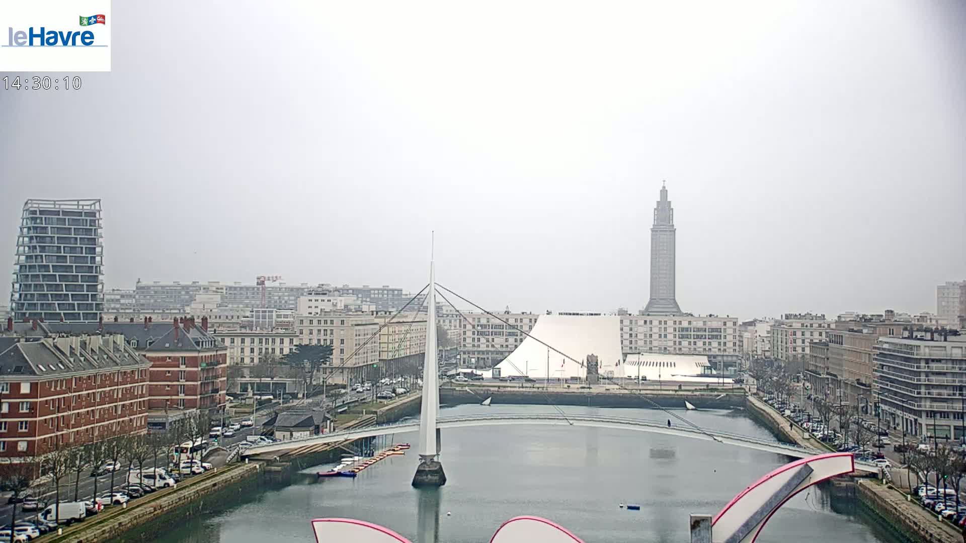 A city harbor scene on an overcast day shows a white pedestrian bridge arching over the water, with buildings, including a tall tower, lining the waterfront.