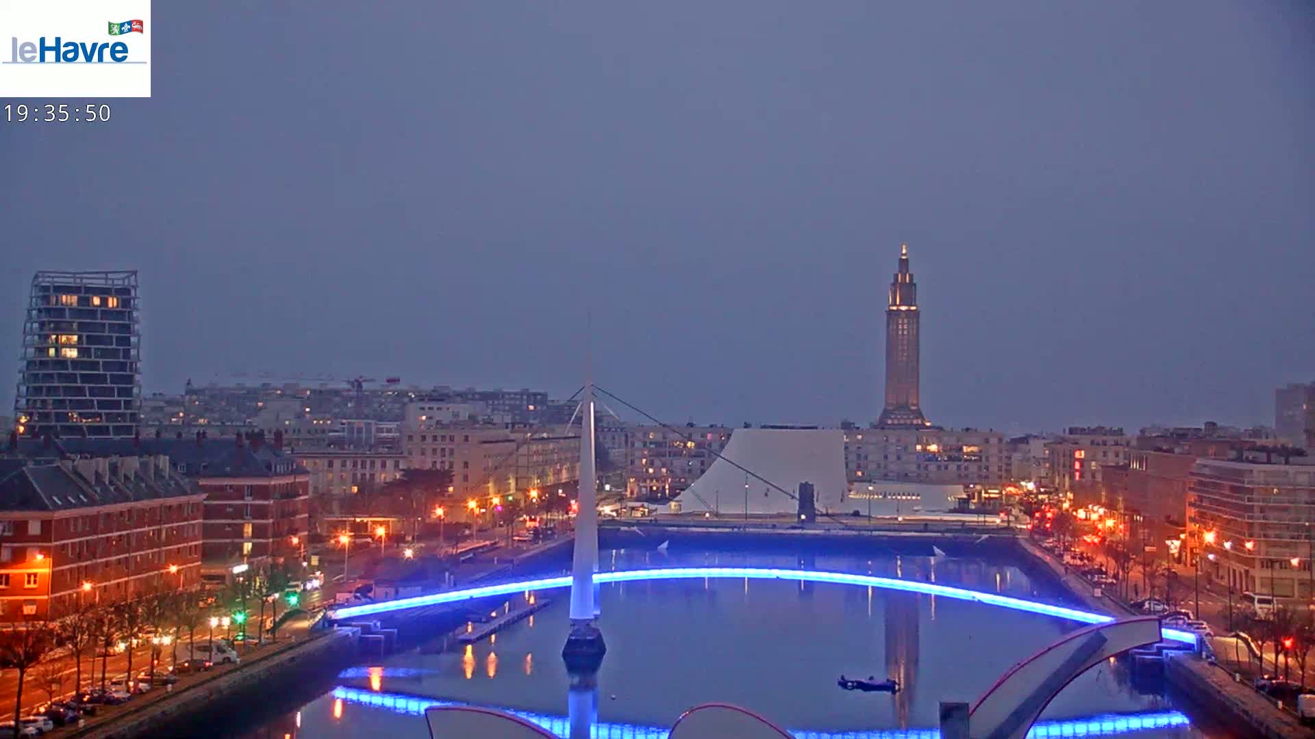A city at night with a body of water reflecting blue lights from a bridge and buildings, under a twilight sky.
