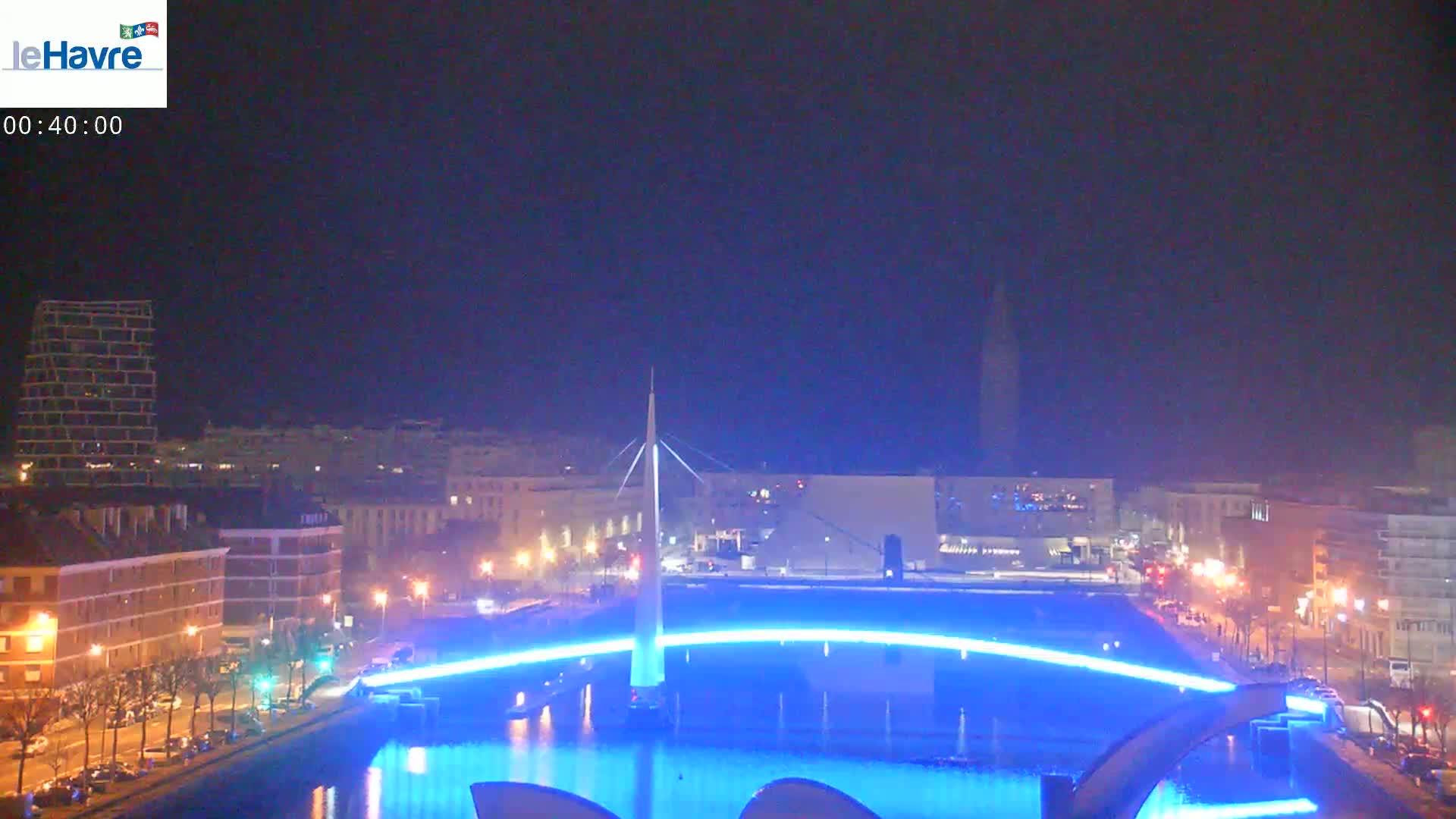A nighttime cityscape is shown with a bridge illuminated in blue light over a body of water.